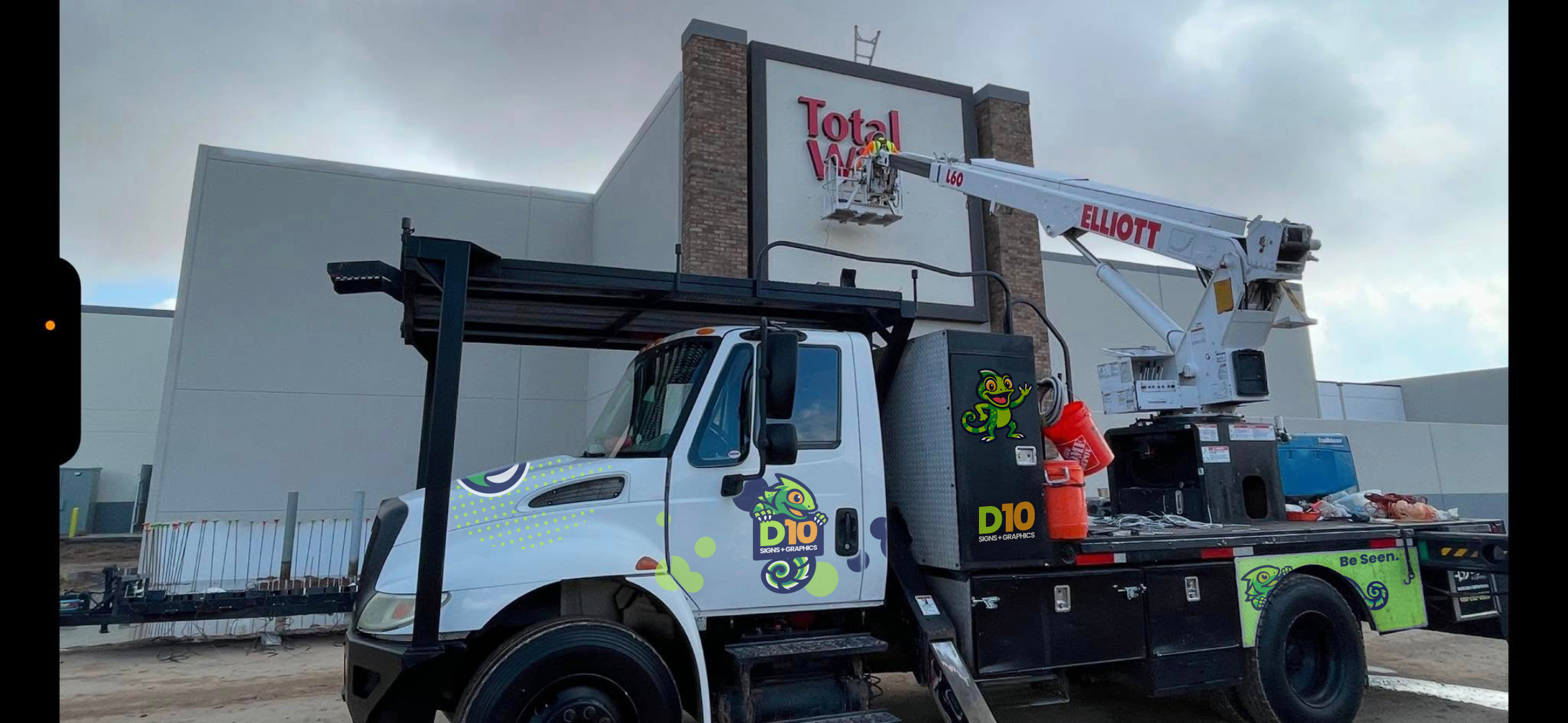 A utility truck with a crane lifting workers to install or repair a sign outside a building. The truck has colorful branding, including a cartoon lizard and the words "D10 Signs & Graphics." The building has a large sign that reads "Total W" on the upper part of the facade, with the workers in a bucket attached to the crane.