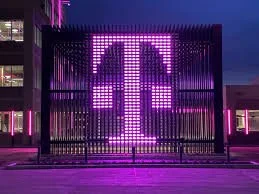 Exterior view of a building at night with a large illuminated pink and purple T-Mobile logo displayed on a fence or screen.