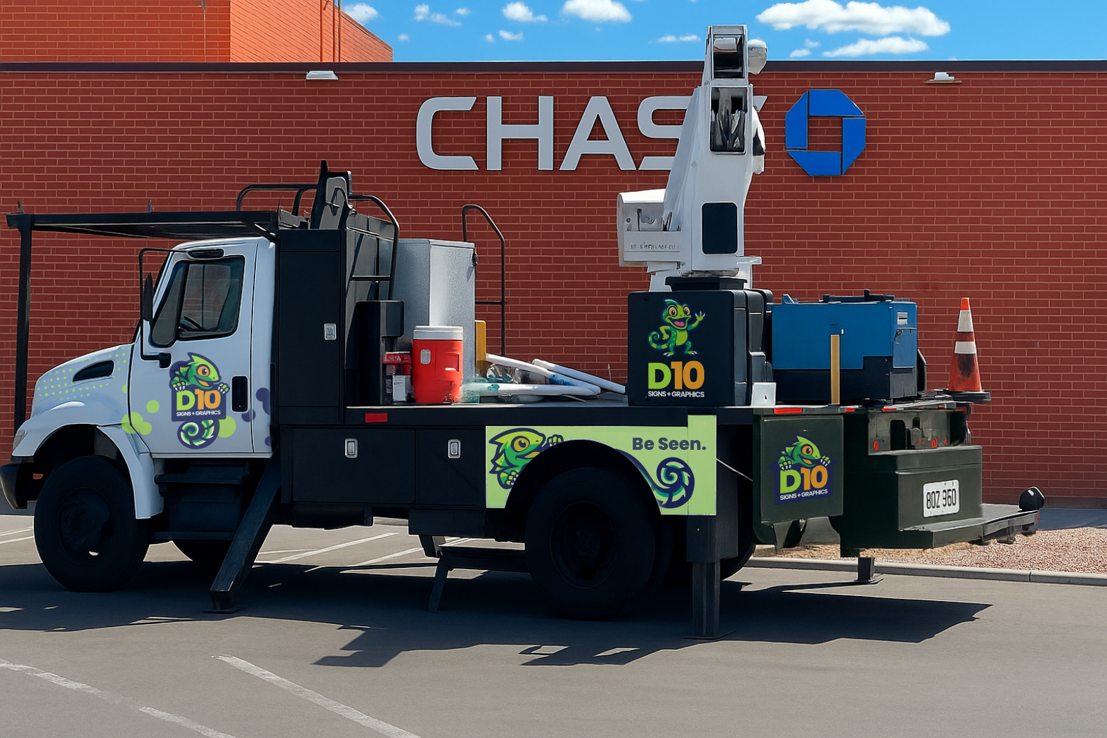 A utility truck with colorful branding parked in front of a red brick Chase Bank building. The truck is equipped with a bucket lift and has graphics of a chameleon and the text "D10 Signs + Graphics" and "Be Seen."