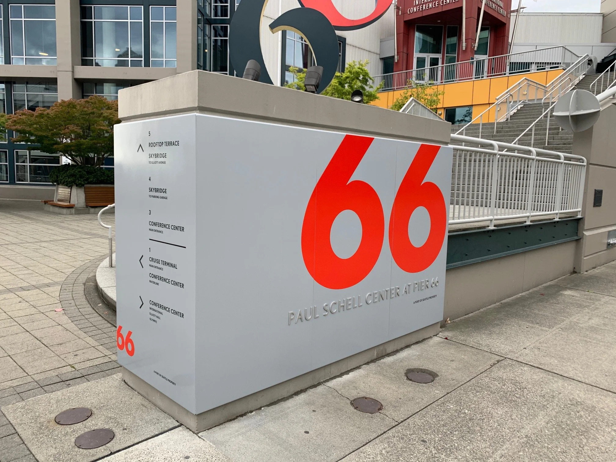 Sign at Pier 66 displaying the address '66' in large red font with the text 'Paul Schell Center at Pier 66' beneath it. The sign also features a navigation guide on the left side indicating locations of the rooftop terrace, Skybridge, conference center, cruise terminal, and main entrance. The background includes staircases, a building, and outdoor urban elements.