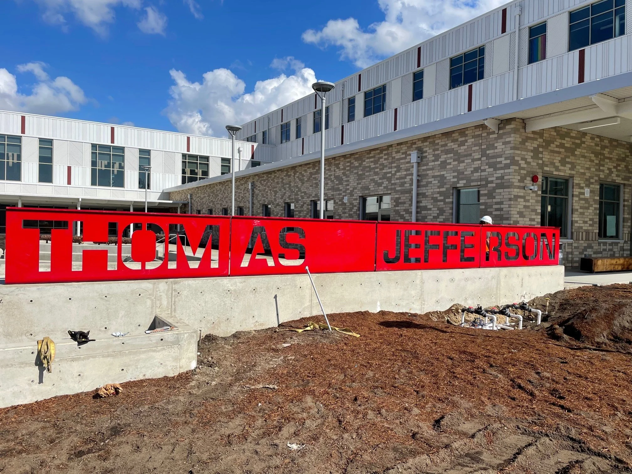Red sign with black cut-out letters spelling 'THOMAS' and 'JEFFERSON' in front of a modern brick and white building under a partly cloudy sky.