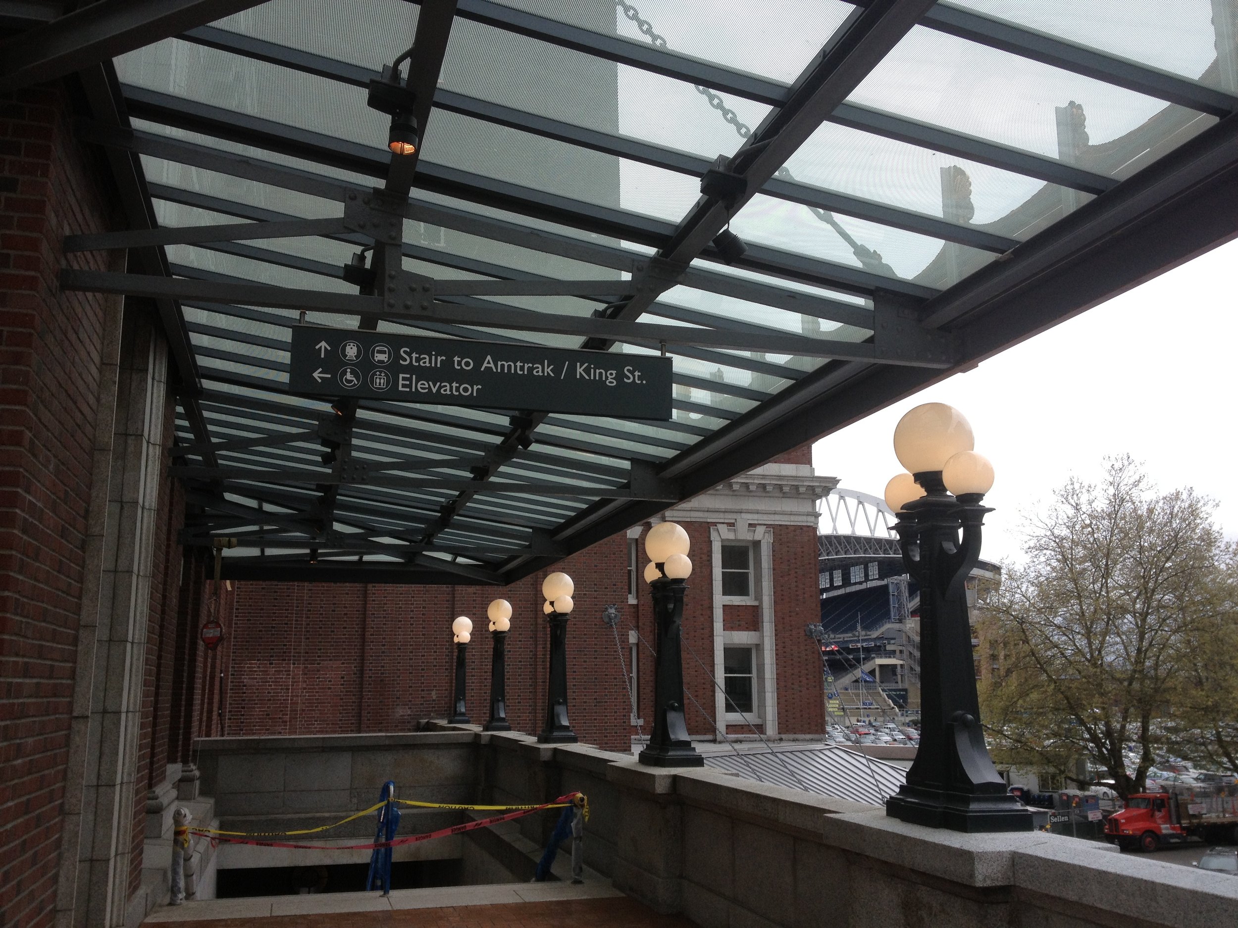 Outdoor area with brick walls, glass roof, and decorative black lamp posts with white globe lights. A sign indicates stairs to Amtrak and King Street, and the elevator. Parking lot with trees and trucks visible in the background.