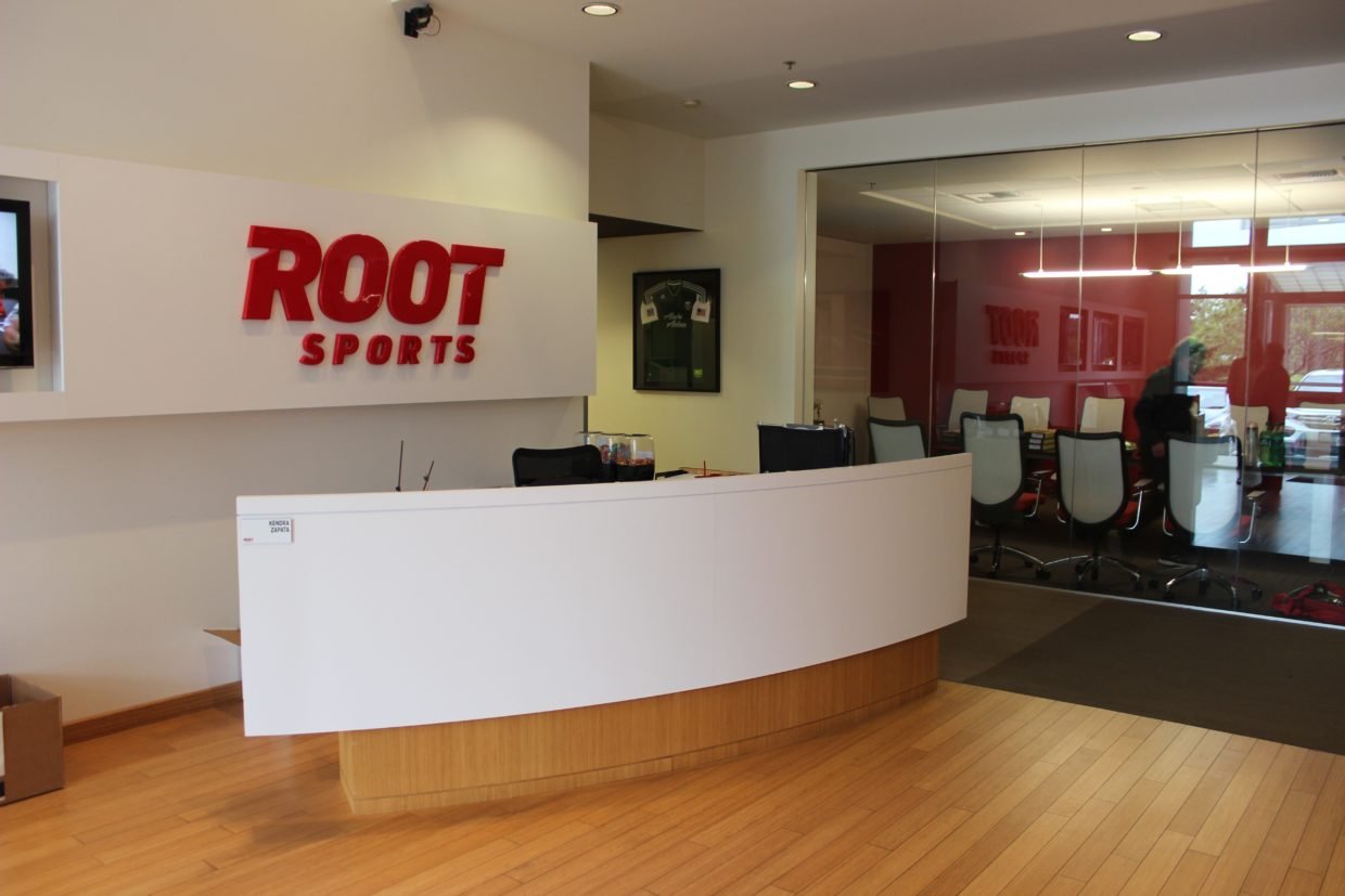 Interior of a ROOT Sports office or studio with a white reception desk, a wall-mounted ROOT Sports sign in red, a framed sports jersey, a glass-walled conference room with chairs, and a wood floor.