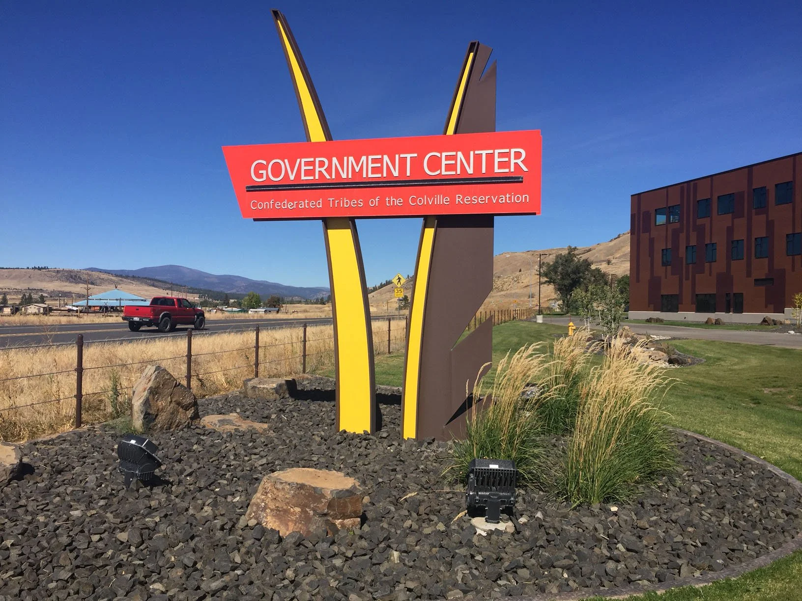 A roadside sign for the Government Center of the Confederated Tribes of the Colville Reservation, with hills, a building, and a red truck in the background.