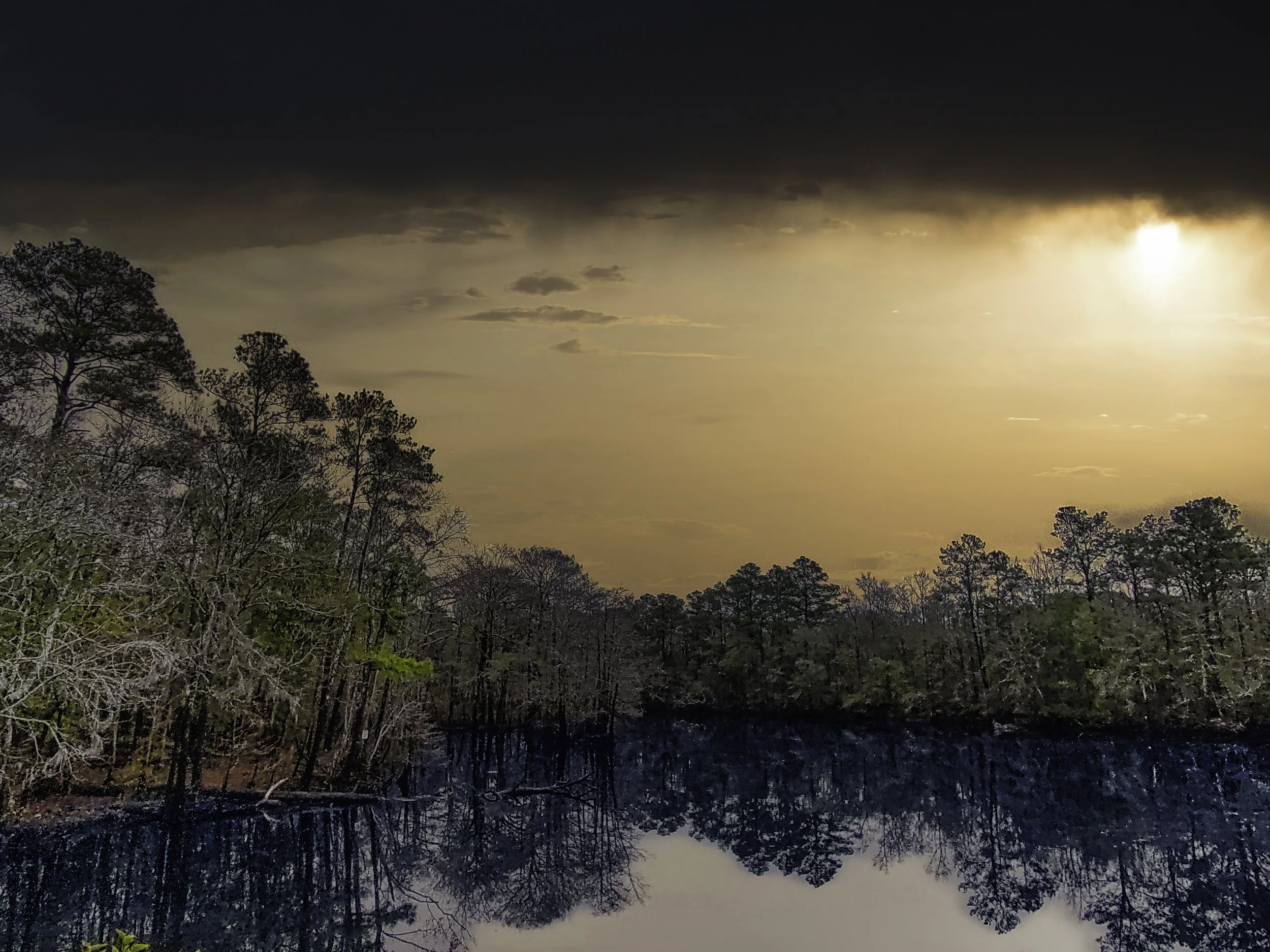 A scenic view of trees reflected in a calm body of water during sunset with dark clouds and the sunlight peeking through.