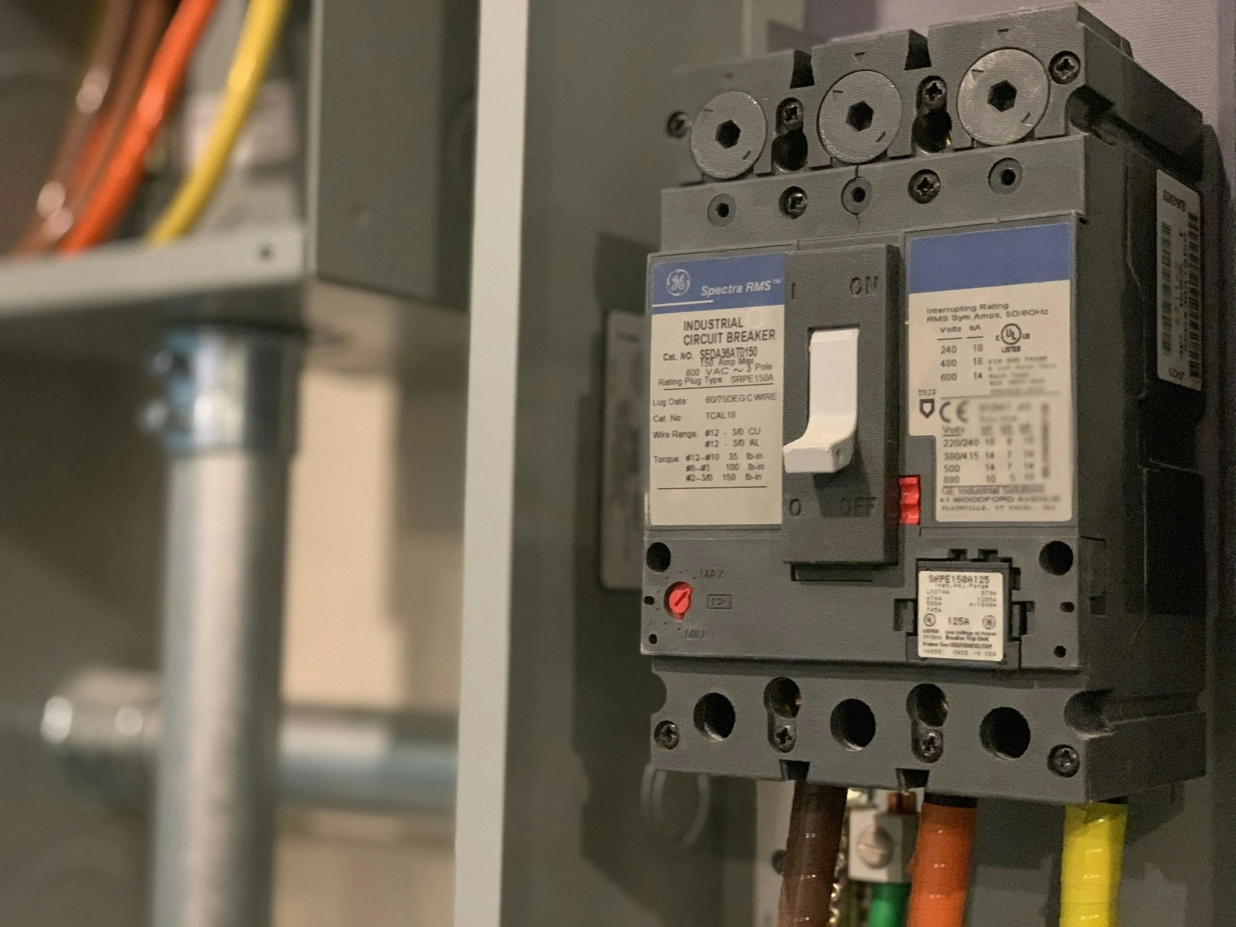 A gray industrial circuit breaker with labels, mounted on a gray electrical panel with colored wires connected at the top and bottom.