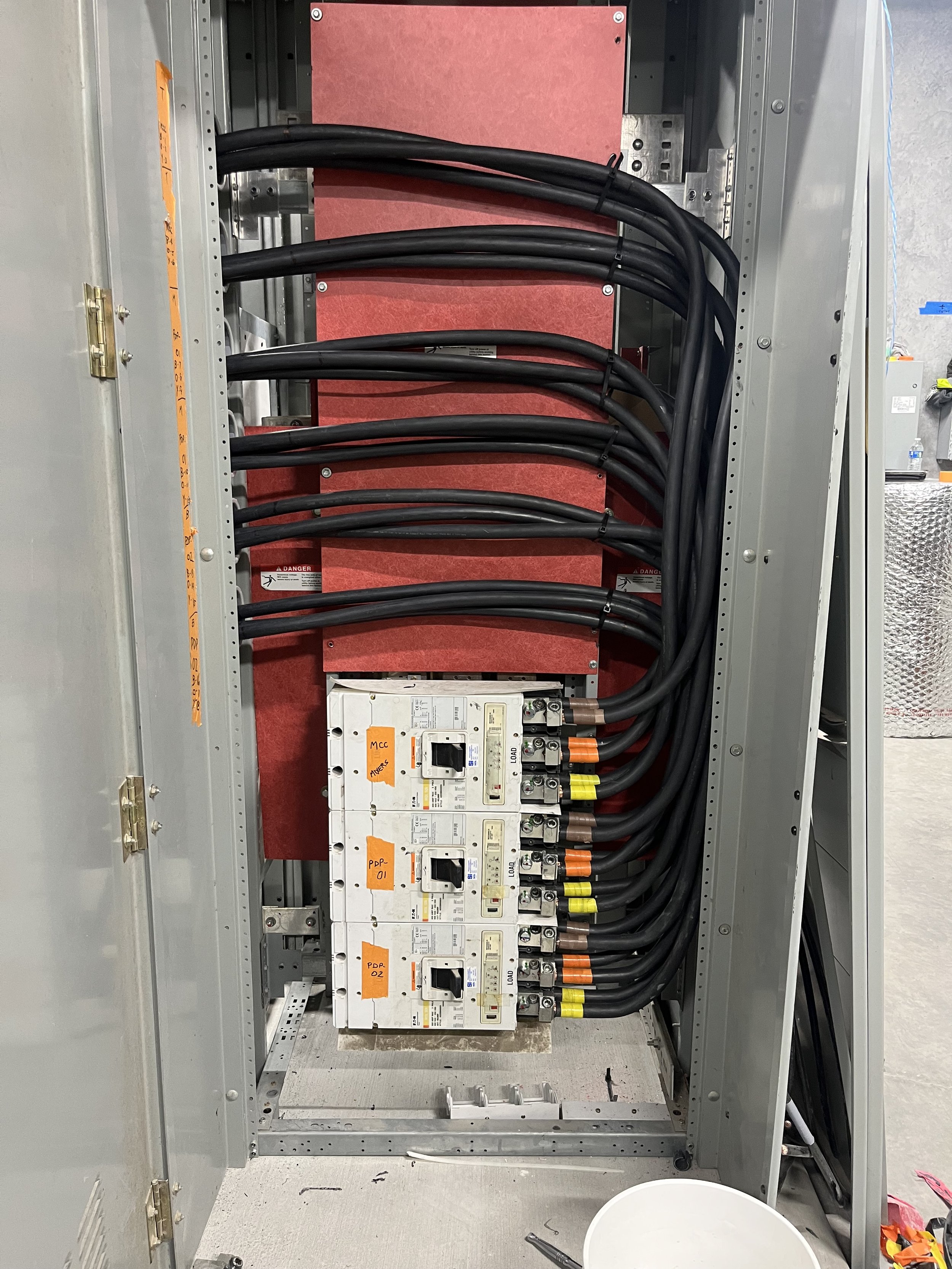 Electrical cabinet with black cables connected to circuit breakers labeled MCC and PDR-01, inside a metal enclosure with a red background panel.