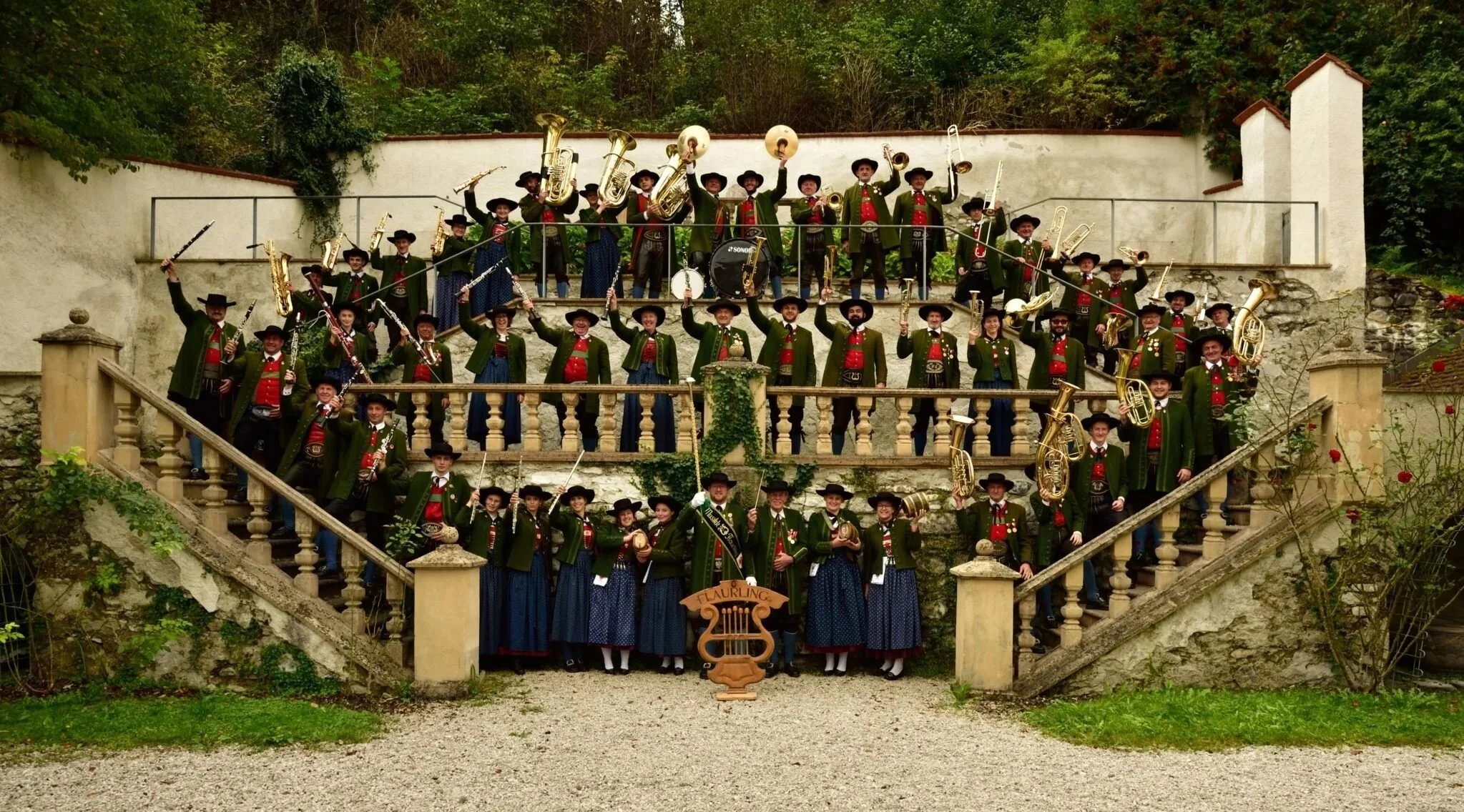 Gruppe von Menschen in traditionellen Trachten, die eine Blaskapelle bilden, auf einer Treppe im Freien, umgeben von Natur, mit verschiedenen Instrumenten.