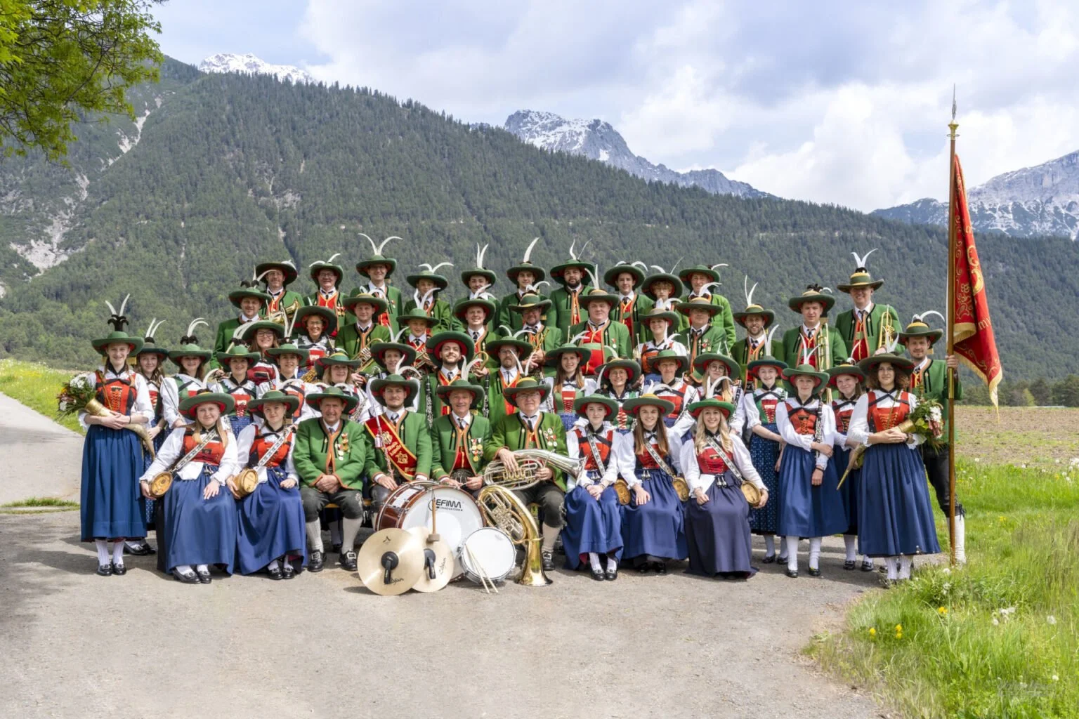 Gruppenfoto von Musikern in traditionellen bayerischen Trachten mit Musikinstrumenten vor einer Bergkulisse.