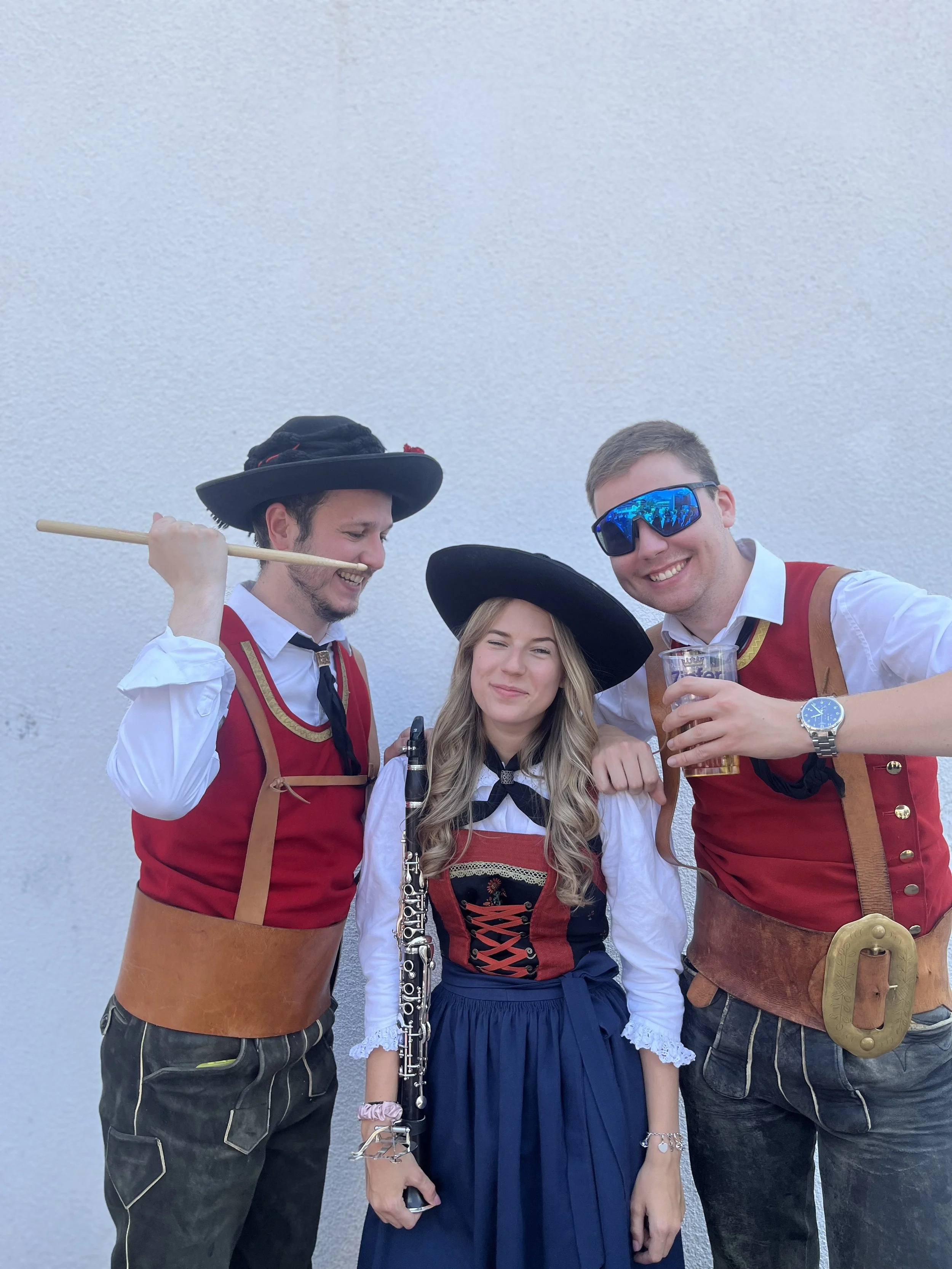 Drei junge Menschen in Trachten feiern zusammen, einer mit einem Keyboard, einer mit Bier, alle lachen vor einer weißen Wand.