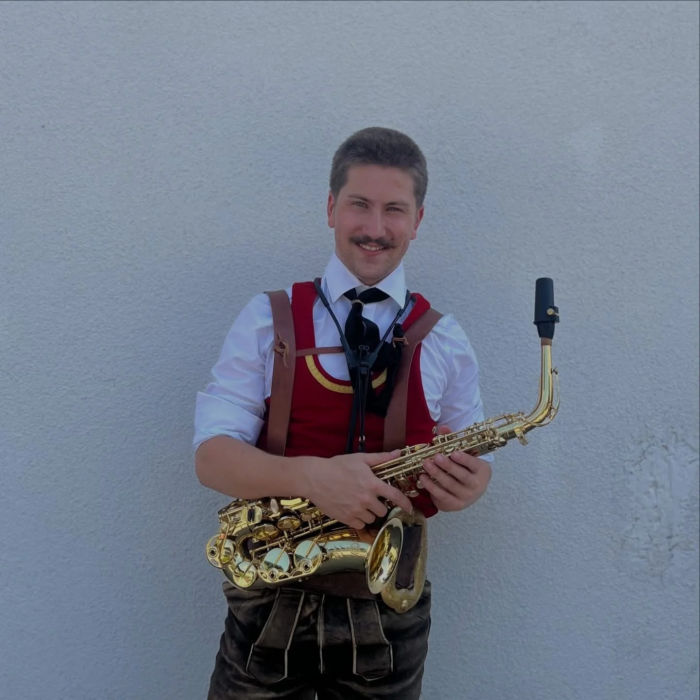 Ein Mann in traditioneller Tracht mit Saxofon vor einer weißen Wand, lächelnd in die Kamera schauend.