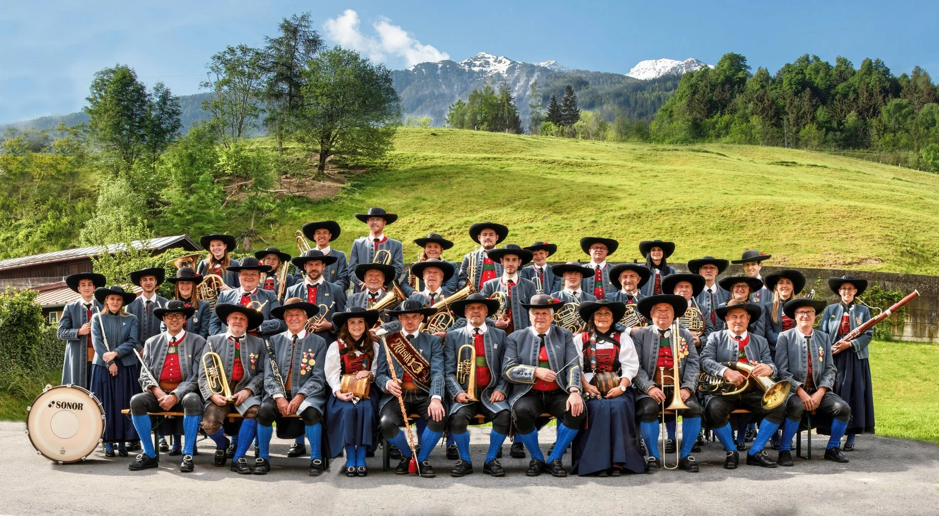 Eine Gruppe von Musikern in traditionellen Trachten steht im Freien vor einer grünen Landschaft mit Hügeln und Bergen
