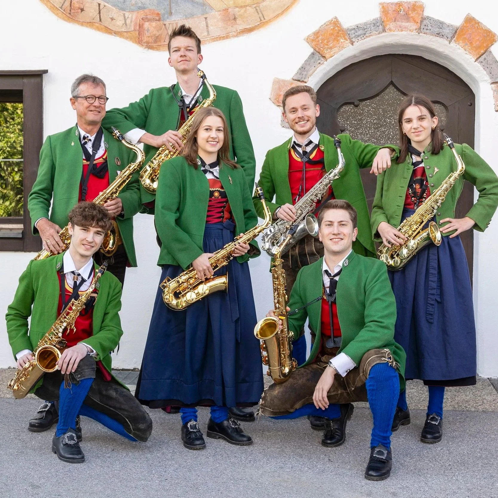 Eine Gruppe von acht jungen Musikern in traditionellen Trachten mit grünen Jacken und Dirndln, die ihre Saxophon-Instrumente halten, vor einem weißen Gebäude mit Steingiebel und einer kleinen Tür.