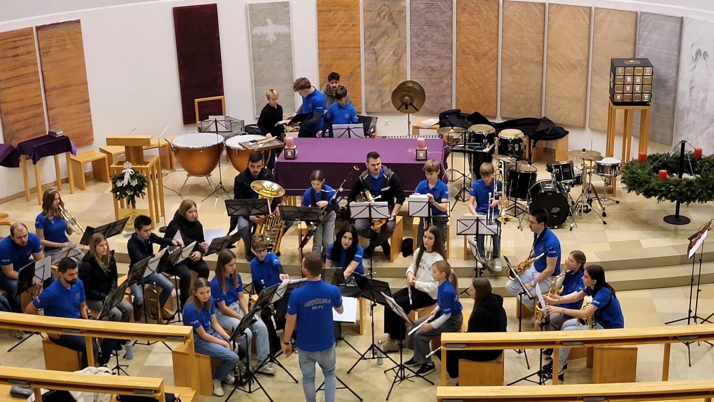 Jugendblasorchester proben in einem mehrstufigen Konzertsaal, mit Holzplatten an den Wänden, Stühlen, Notenständern und verschiedenen Musikinstrumenten wie Trompeten, Saxophonen, Trommeln und einer Pauke.