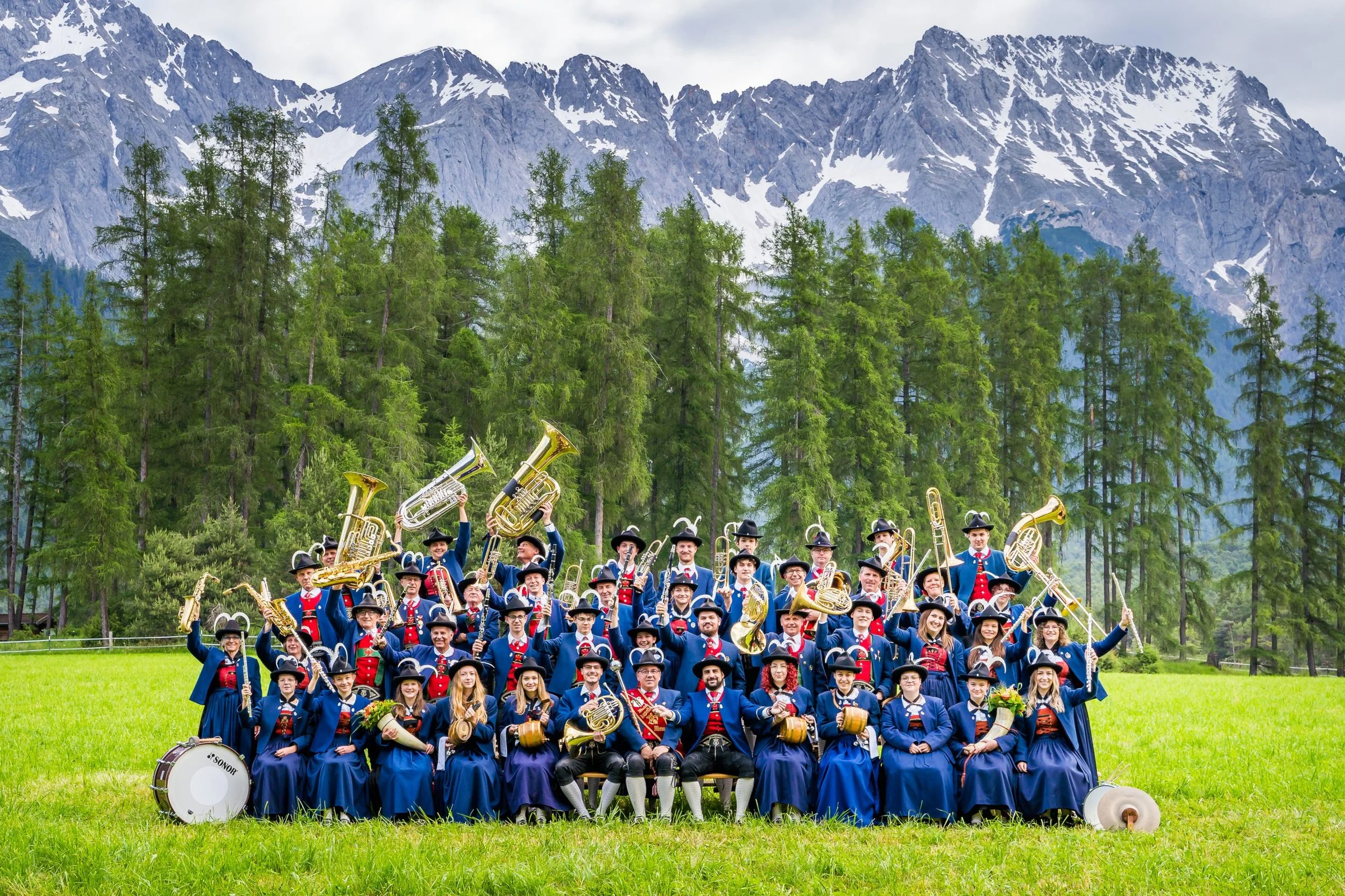 Gruppe von Menschen in traditioneller deutscher Tracht mit Musikern und Instrumenten, im Freien vor einer grünen Wiese, Tannenbäume und Berge im Hintergrund.