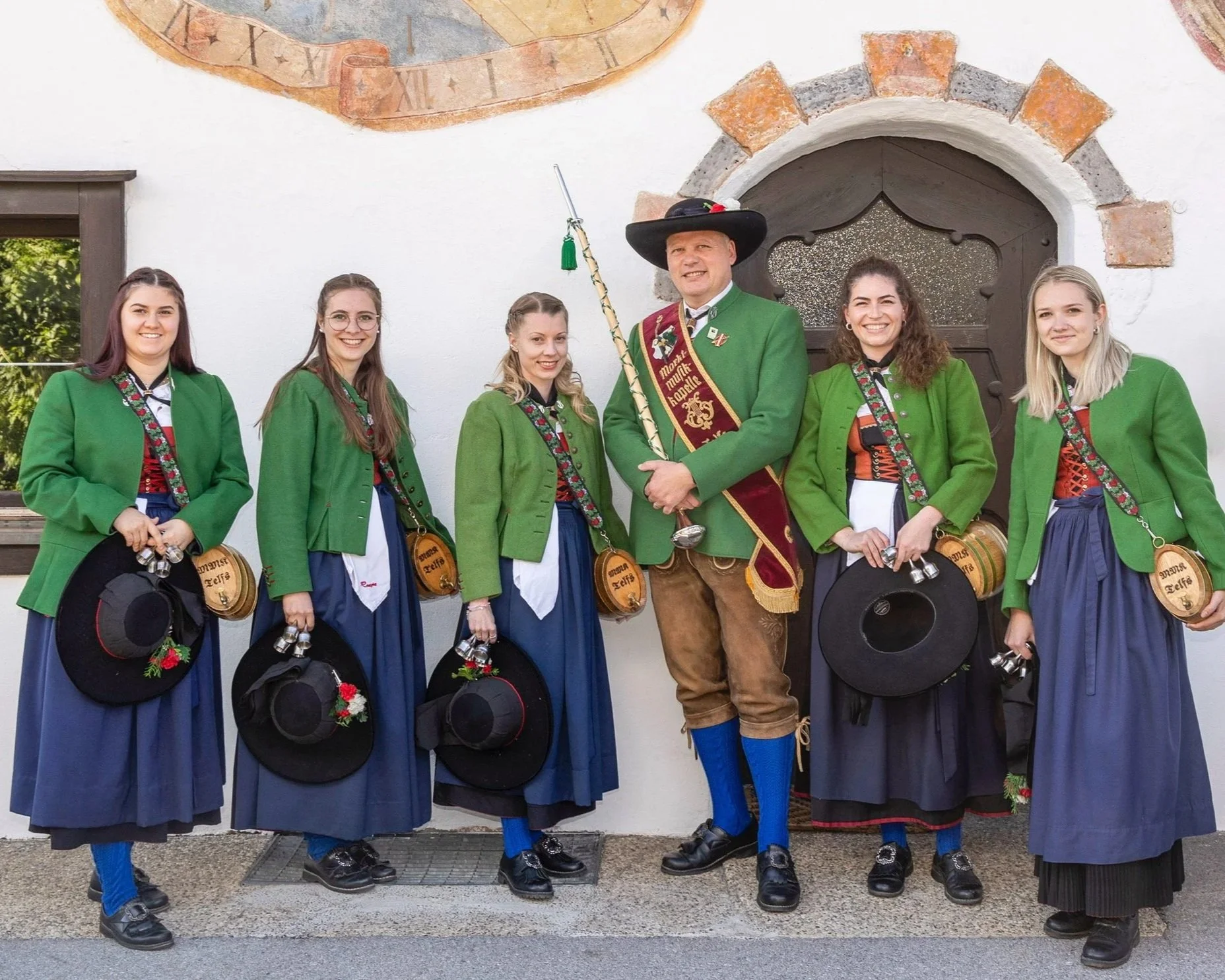 Gruppe von Menschen in traditionellen Trachten, die vor einer weißen Wand mit einer historischen Uhr posieren.
