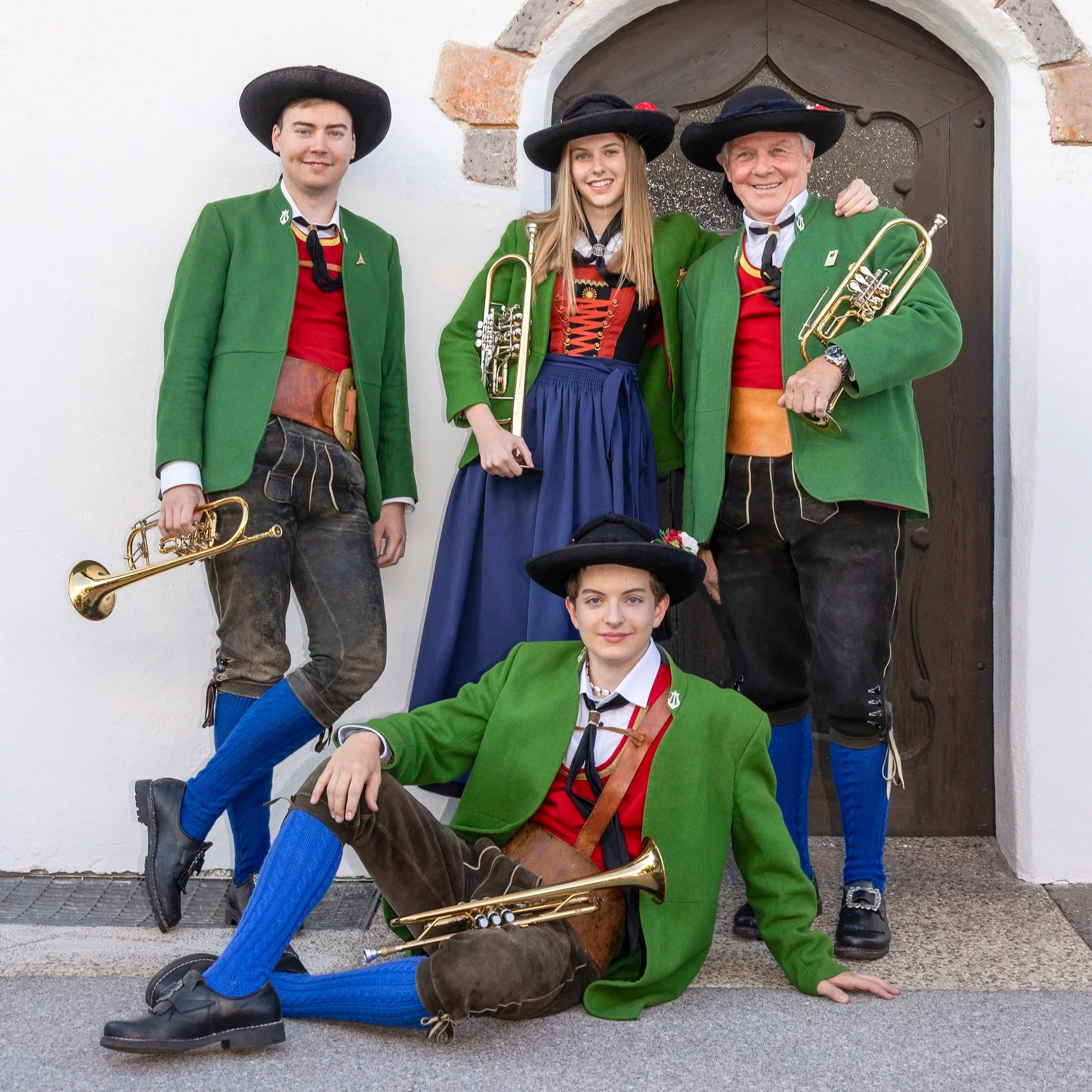 Fünf Menschen in traditionellen bayerischen Trachten, mit Blaskapelleninstrumenten vor einer Holztür.