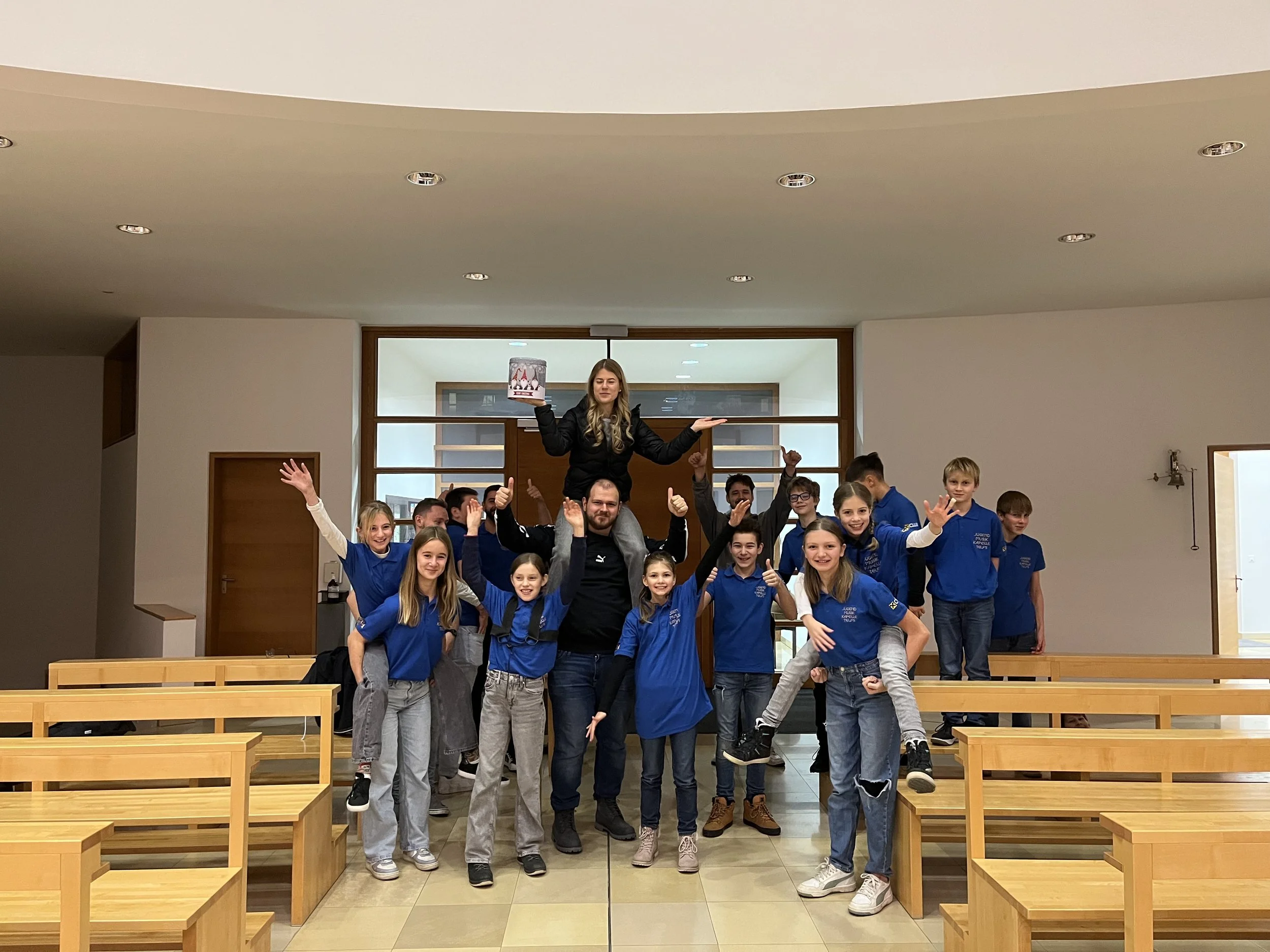 Gruppenfoto von Jugendlichen in blauen T-Shirts, die im Raum feiern, mit einer Frau auf den Schultern, die ein Schild hält.