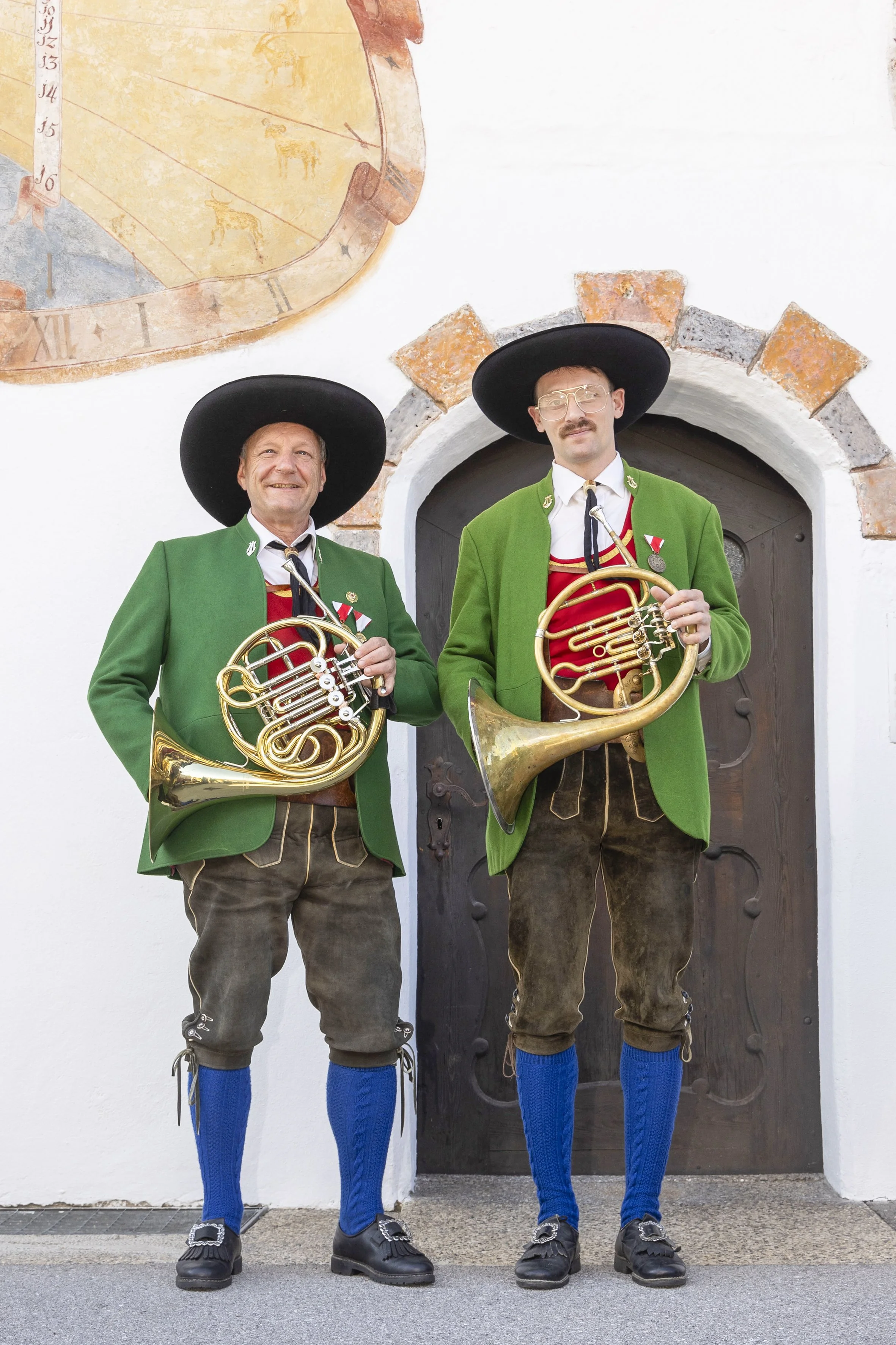 Zwei Männer in traditioneller bayerischer Kleidung mit grünen Jacken, knielangen Lederhosen, blauen Socken, schwarzen Schuhen und großen schwarzen Hüten stehen vor einer Holztür und halten Musikinstrumente, wahrscheinlich Alphörner.