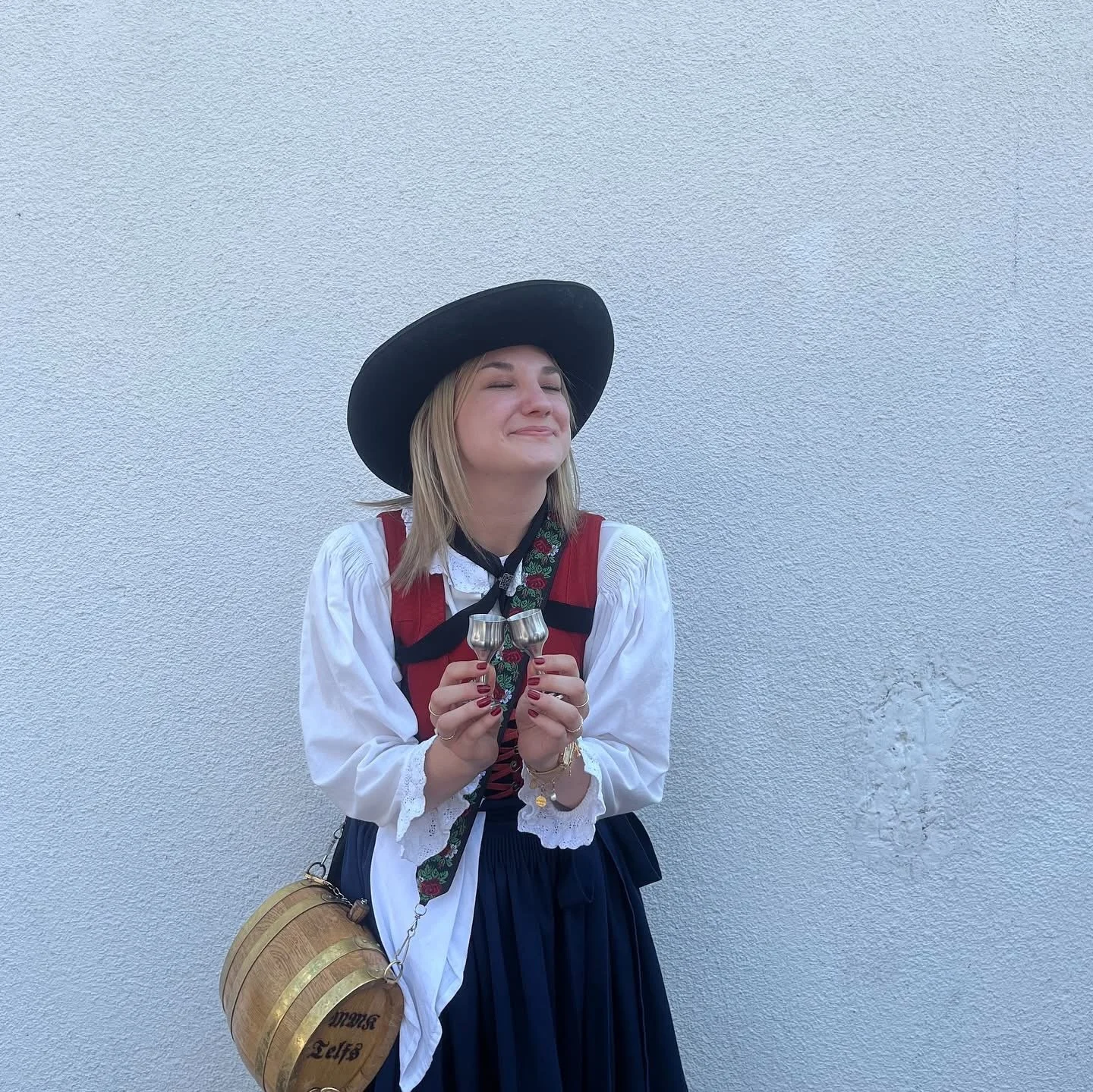 Junge Frau in traditioneller bayerischer Tracht, trägt einen schwarzen Hut, hält zwei Gläser und steht vor einer weißen Wand.