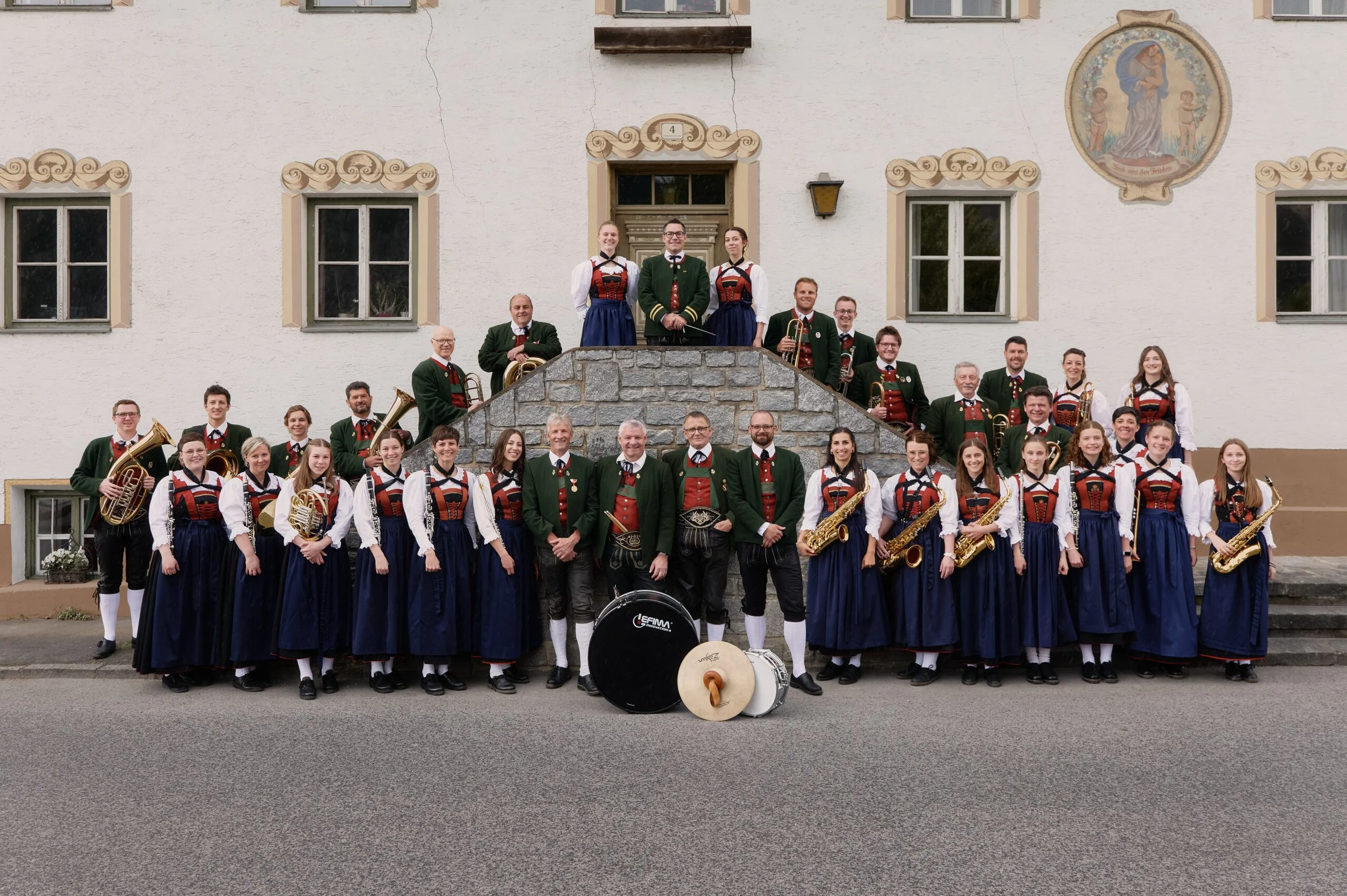Gruppe von Musikern in traditioneller bayerischer Kleidung vor einem Gebäude, einige spielen Instrumente.