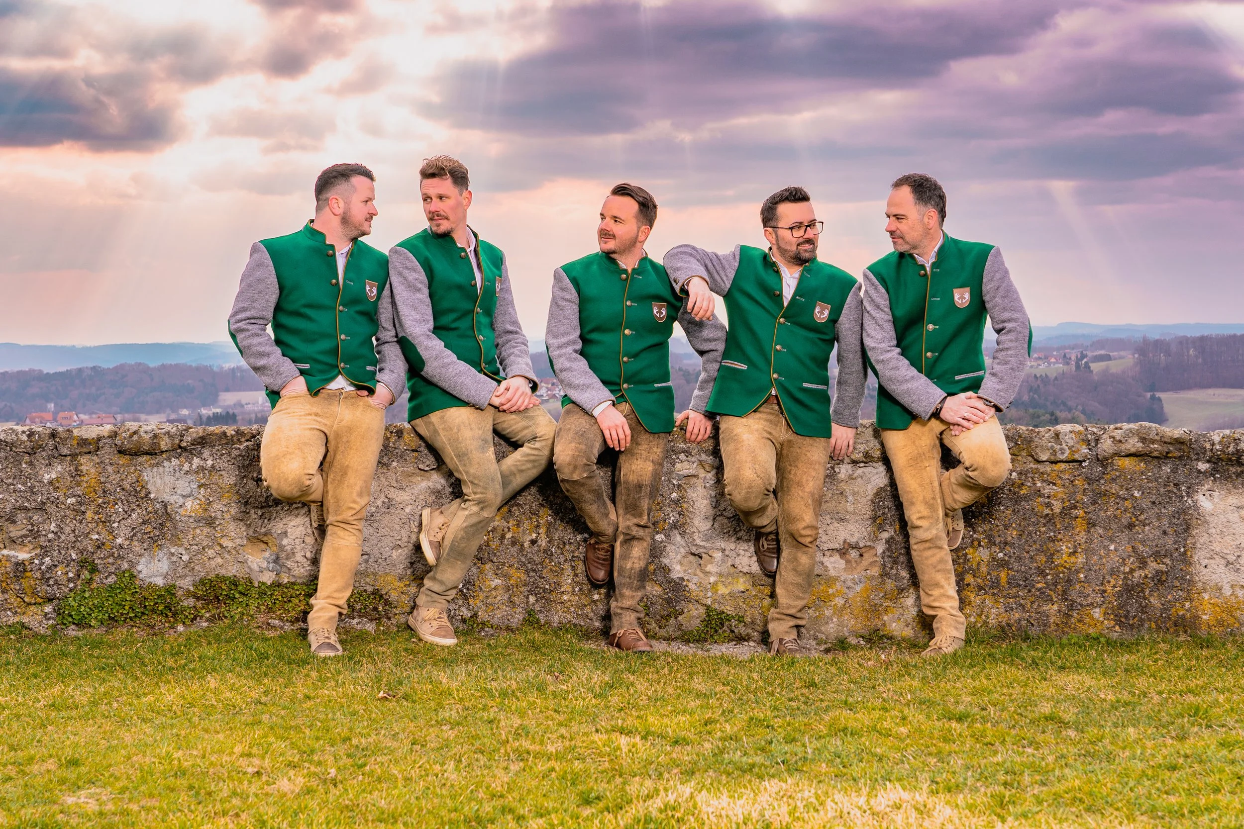 Fünf Männer in traditionellen Trachten stehen vor einer Mauer im Freien mit einer ländlichen Landschaft im Hintergrund bei Sonnenuntergang.