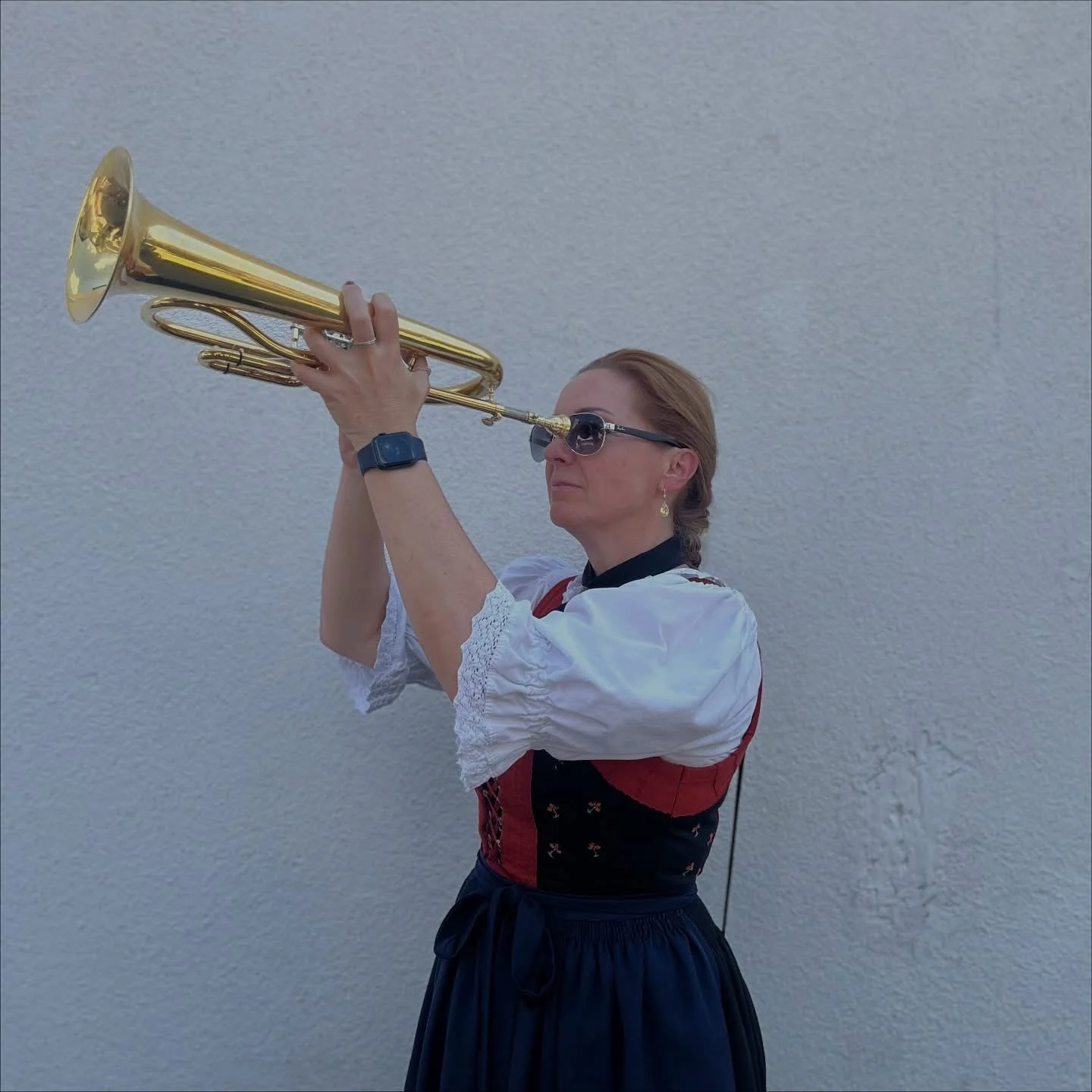 Frau in traditionellen Tracht hält eine Trompete in der Hand und schaut durch eine Sonnenbrille nach oben, vor einer grauen Wand.