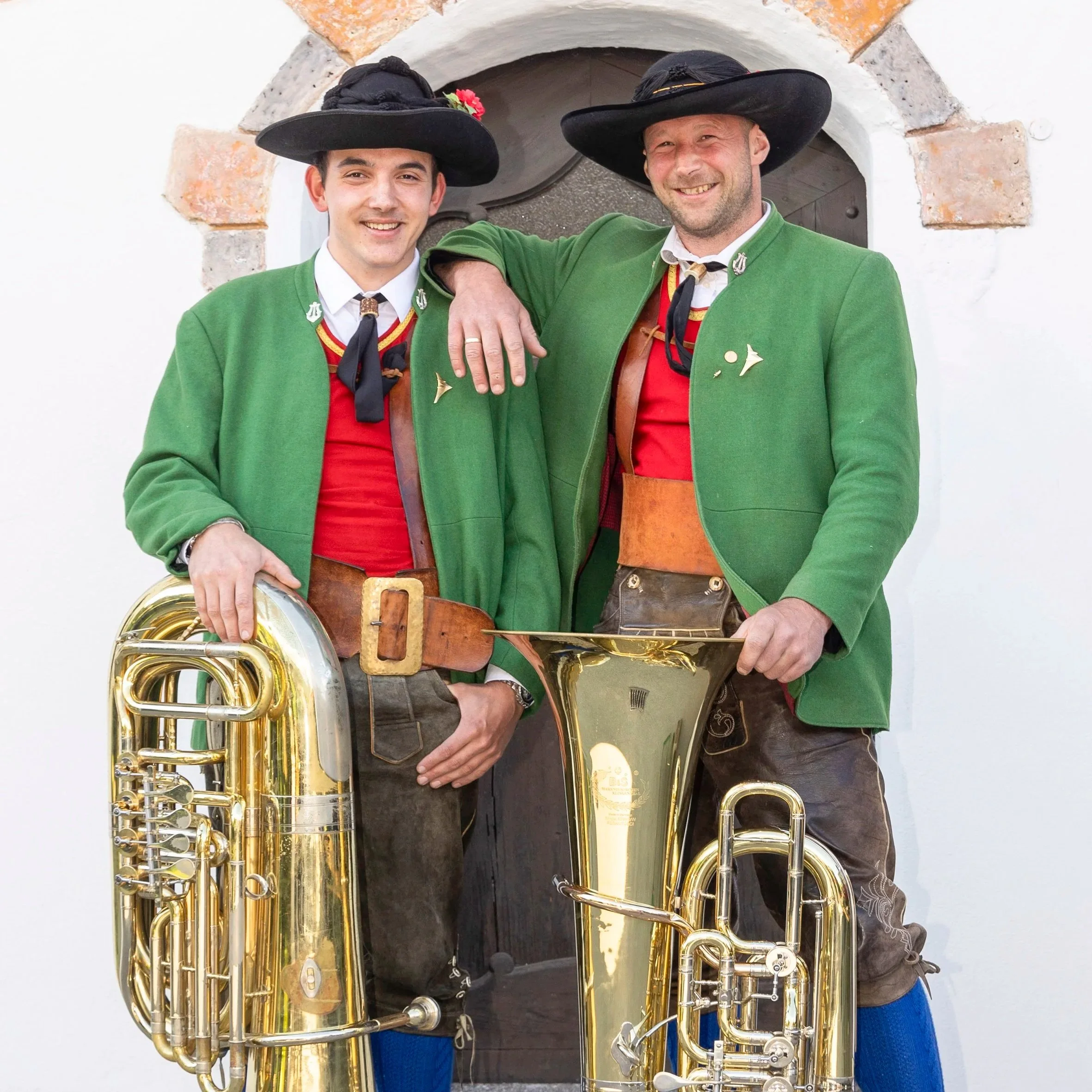 Zwei Männer in traditionellen bayerischen Trachten mit grünen Jacken, schwarzen Hüten und Tuba vor weißem Hintergrund.