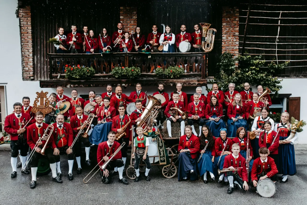 Gruppe von Musikern in traditionellen Trachten, die mit unterschiedlichsten Musikinstrumenten vor einem Gebäude stehen, das mit Holz und Zement gebaut ist.