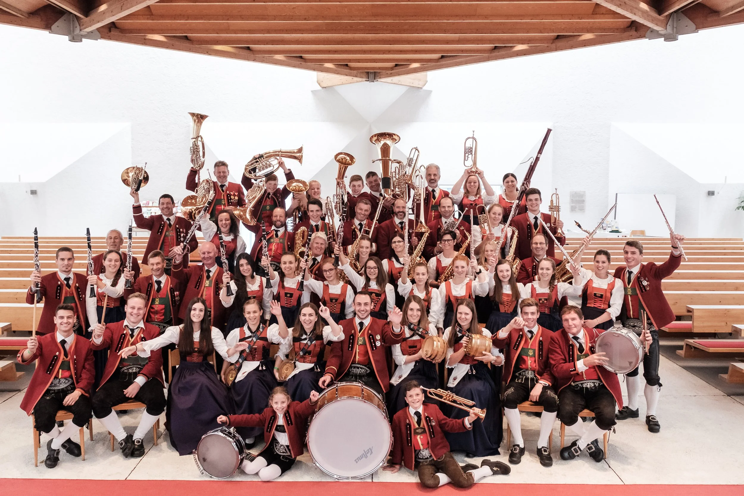 Eine große Gruppe von Musikern in traditionellen trachtähnlichen Kleidung, die in einer Halle zusammenstehen, mit Instrumenten wie Trompeten, Klarinetten, Fagotten, Trommeln und mehr, möglicherweise ein Blasorchester.