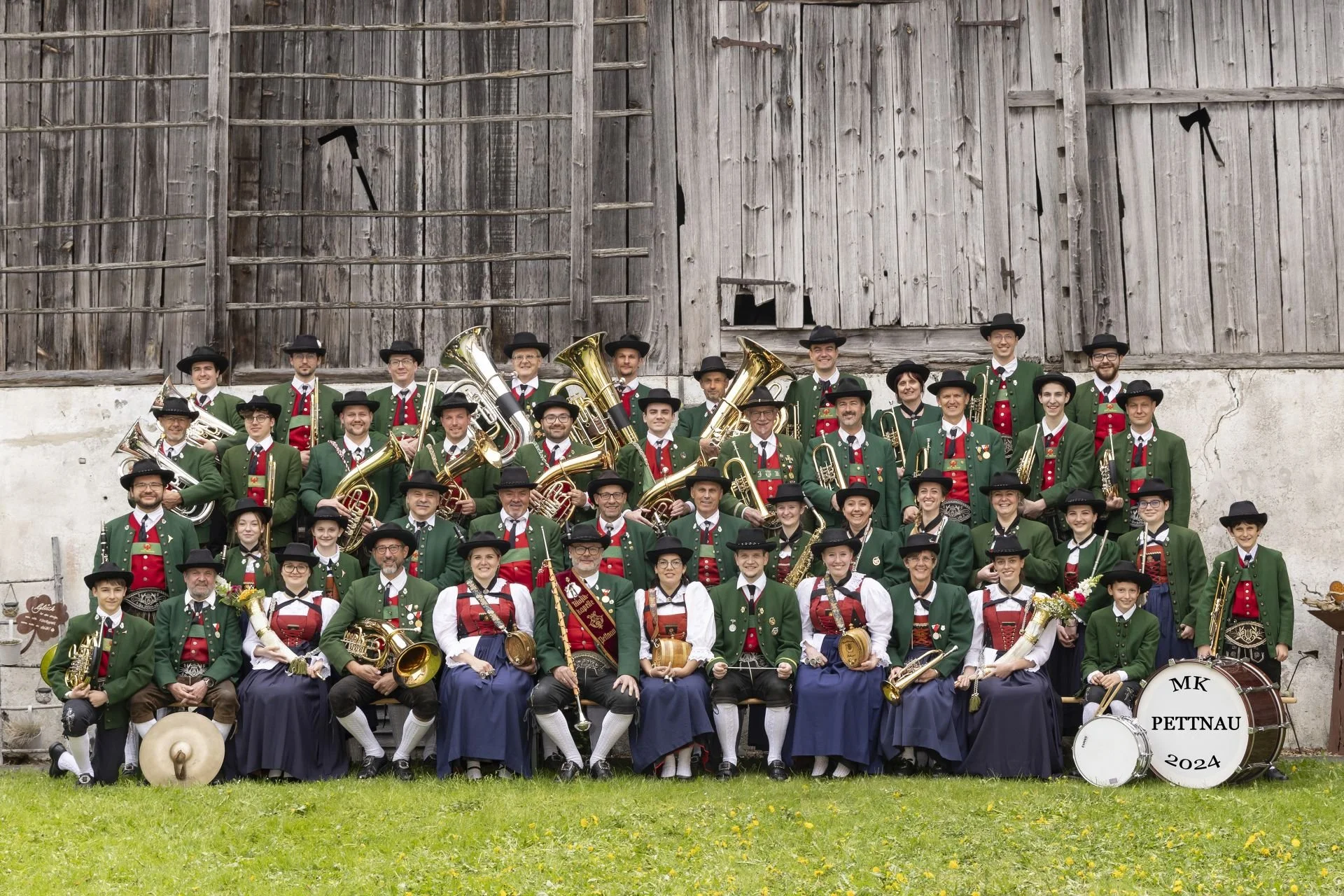 Eine große Gruppe von Menschen in traditionellen Trachten, die eine Blaskapelle bilden, vor einer alten Holzfassade. Sie halten Musikinstrumente wie Trompeten, Tubas, Klarinetten und Schlagzeug, mit einem Schild, das 'MK PETTNAU 2024' schreibt.
