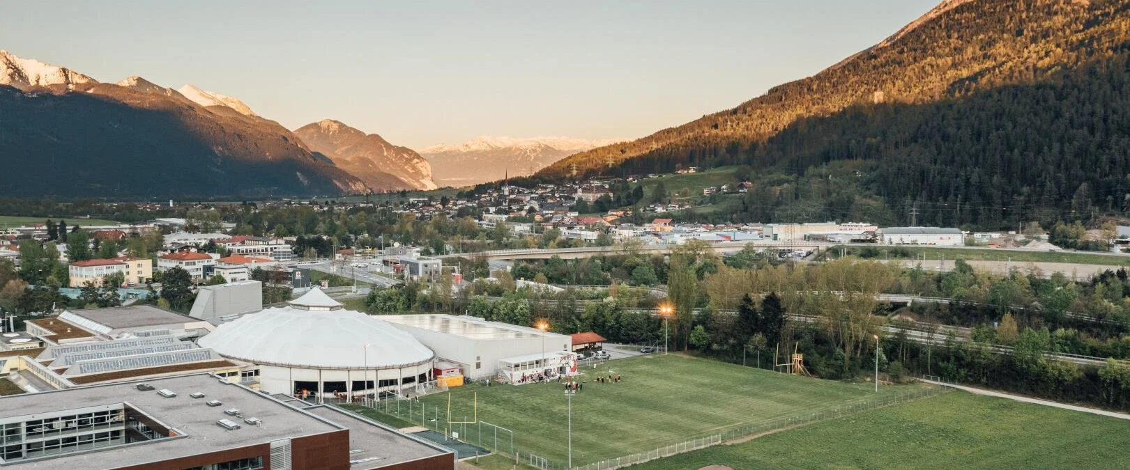 Landschaft eines Tals mit Bergen im Hintergrund und einem Sportfeld im Vordergrund.
