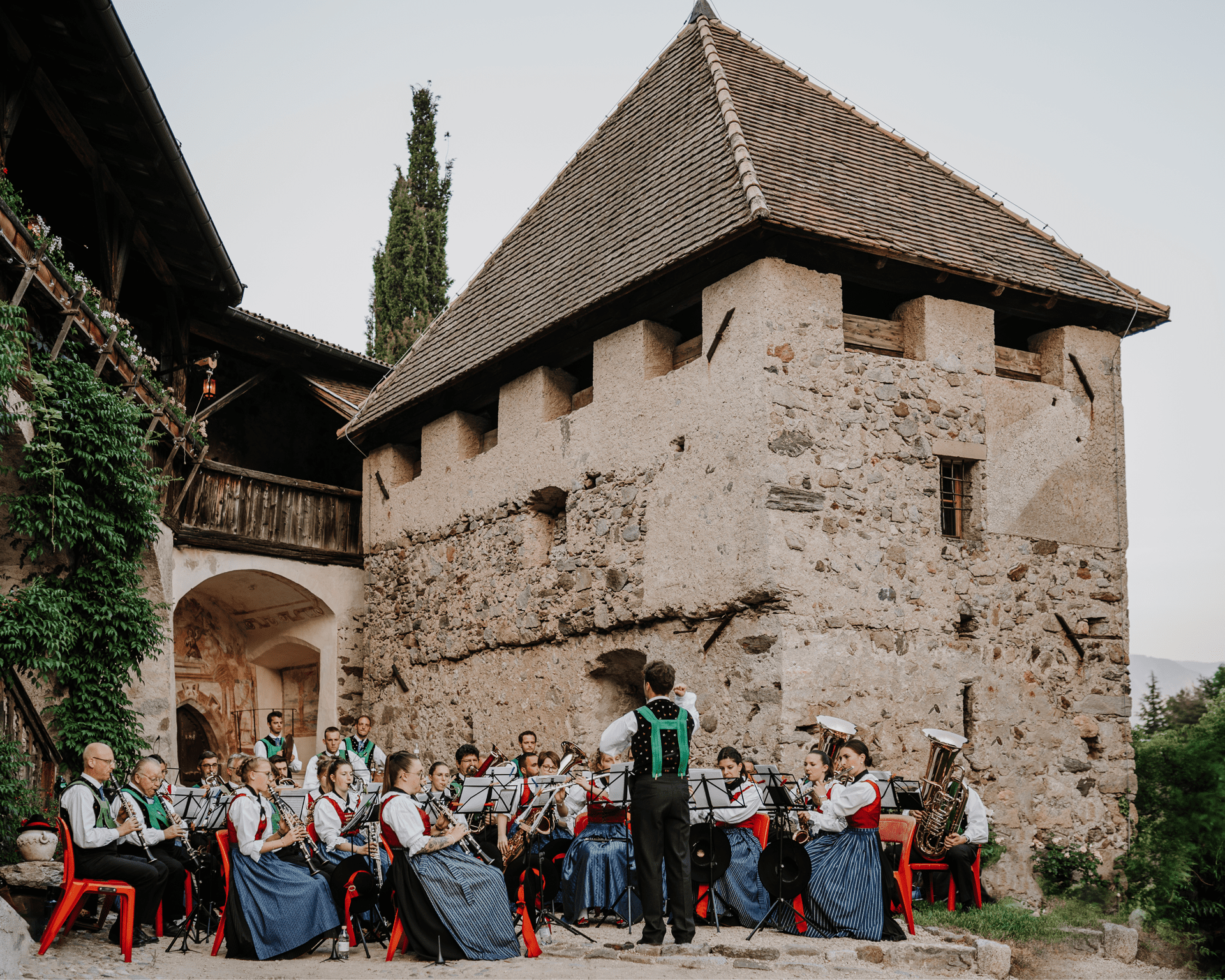 Eine Gruppe von Musikern in traditionellen Trachten spielt vor einem alten Steinturm, während ein Dirigent sie leitet. Im Hintergrund befinden sich alte Gebäudestrukturen und ein dichter grüner Baum.