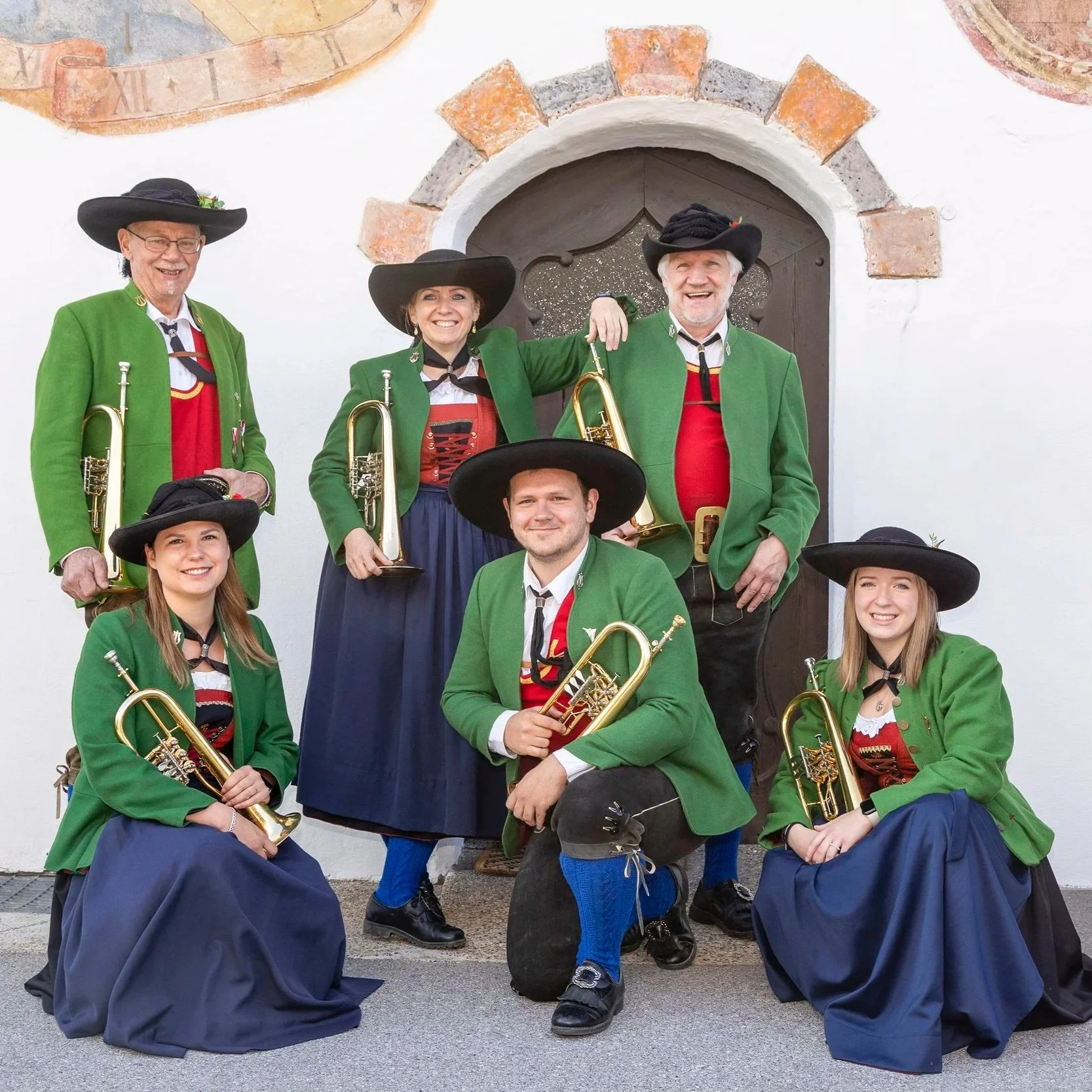 Sieben Musiker in traditionellen bayerischen Trachten mit grünen Jacken und schwarzen Hüten, die eine Blaskapelle bilden, vor einer weißen Wand mit einer Holztür im Hintergrund.