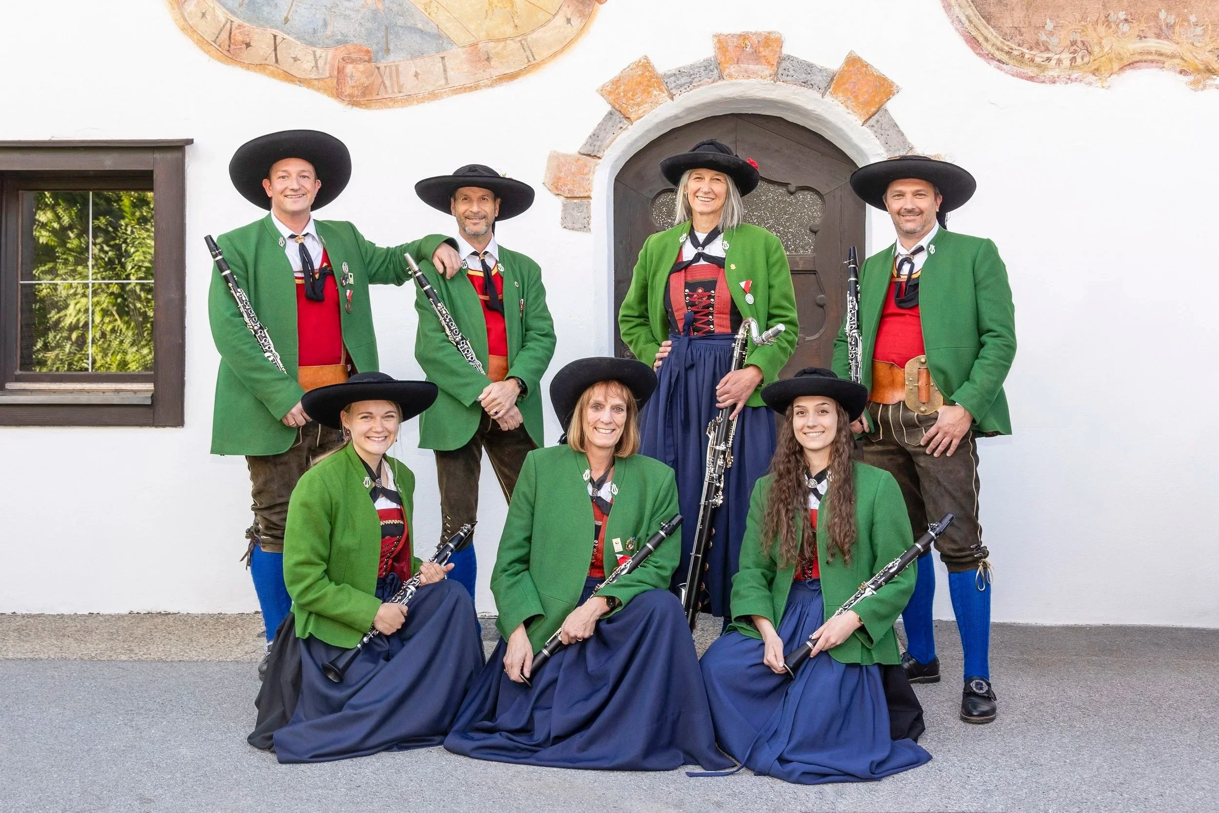 Eine Gruppe von acht Menschen in traditioneller bayerischer Tracht mit grünen Jacken, roten und schwarzen Oberteilen, schwarzen Hüten und Blasinstrumenten, als Blaskapelle vor einer weißen Wand mit einer kleinen Fenster und einem Rundbogen mit Steinrahmen