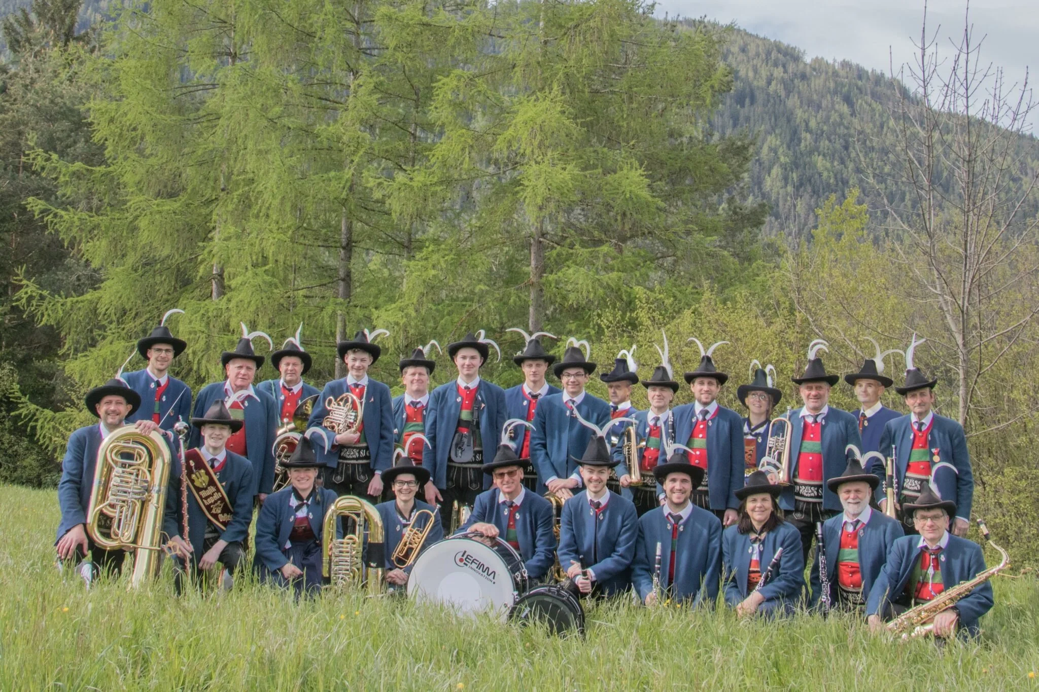 Eine Gruppe von Musikern in traditionellen Trachten mit schwarzen Hüten, die auf einer Wiese im Freien stehen, umgeben von Bäumen und Bergen im Hintergrund, während sie ihre Musikinstrumente halten.