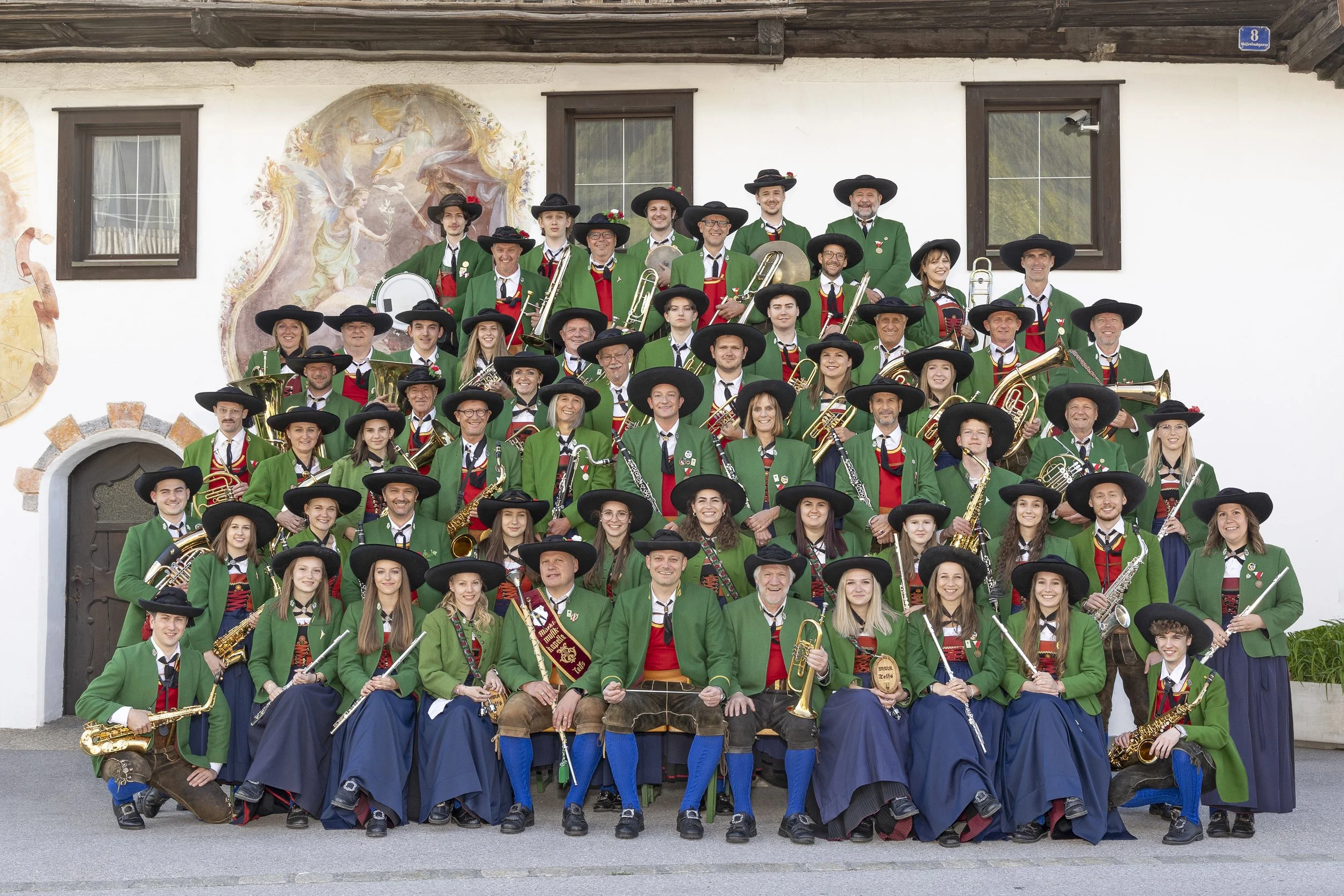 Eine große Gruppe von Musikern in traditionellen bayerischen Trachten mit grünen Jacken, roten Westen und schwarzen Hüten, die vor einem Gebäude stehen, das Wandmalereien zeigt.