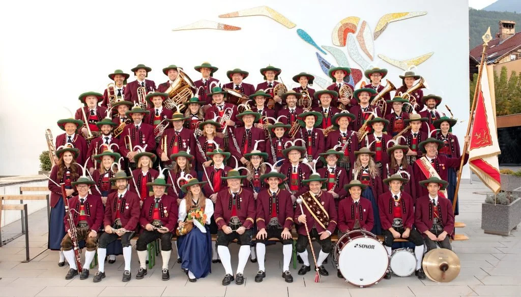 Eine große Gruppe von Männern, Frauen und Kindern in traditionellen bayerischen Trachten mit Musikinstrumenten, vermutlich eine Blaskapelle, steht vor einer weißen Wand mit einem bunten Vogel-Motiv.