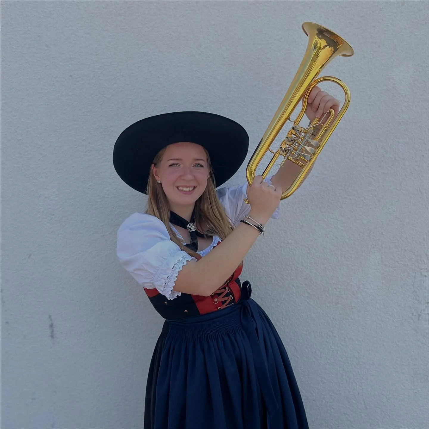 Junge Frau in traditioneller Tracht mit Hut, die eine Trompete hält und lächelt, vor einer weißen Wand.
