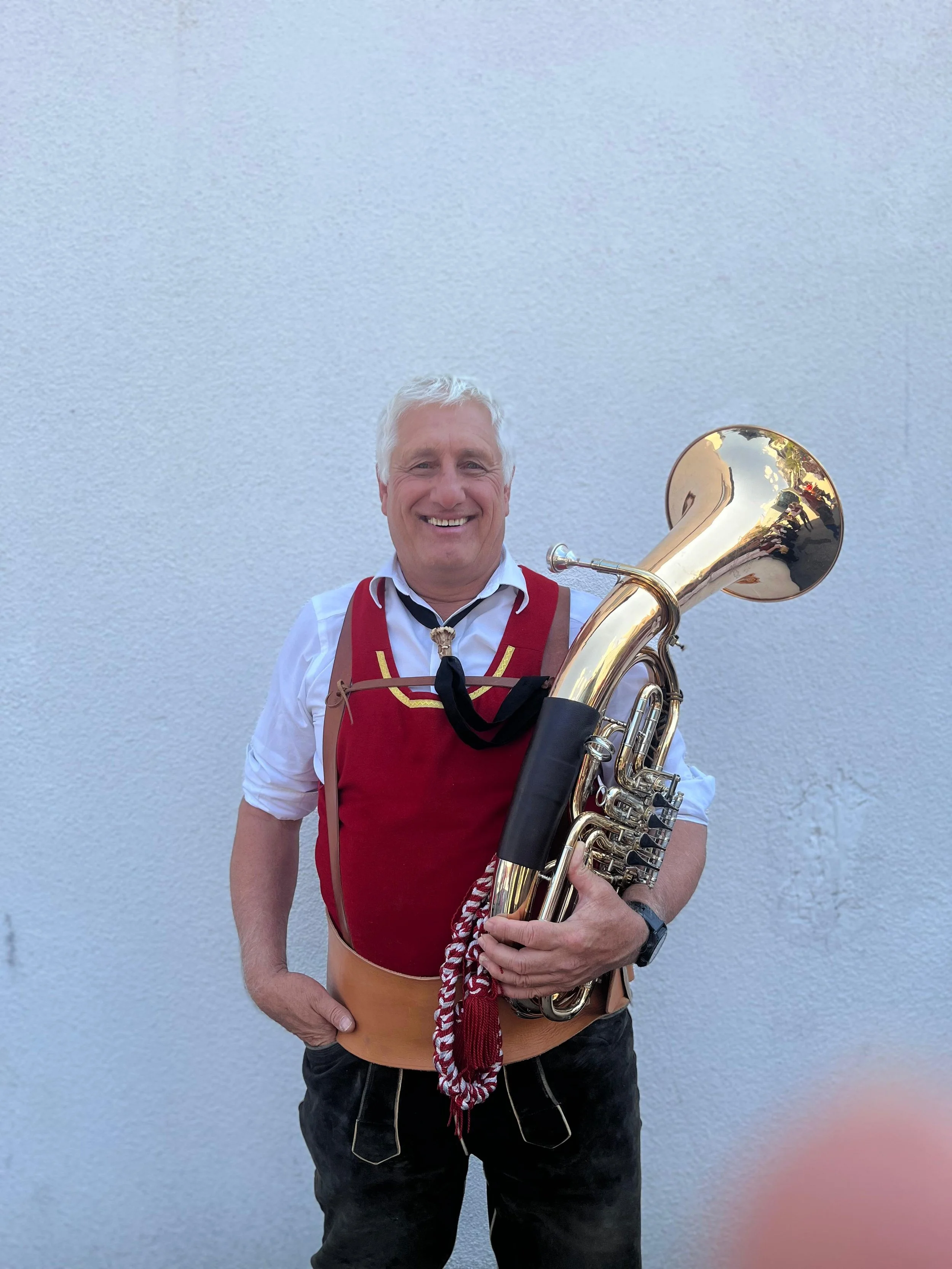 Ein Mann in traditioneller Tracht hält ein Tuba vor einer weißen Wand.