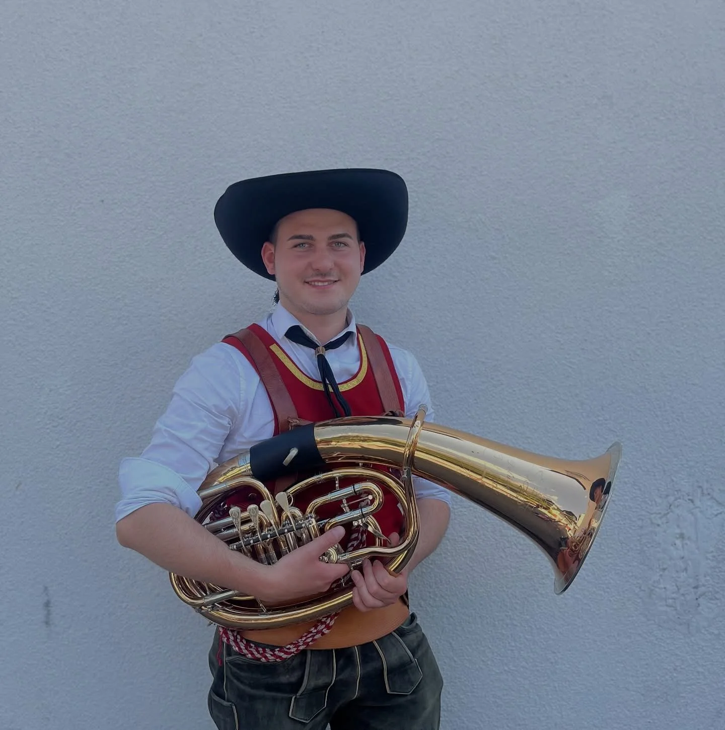 Junge in traditioneller Tracht mit Hut hält ein Saxophon vor einer weißen Wand.