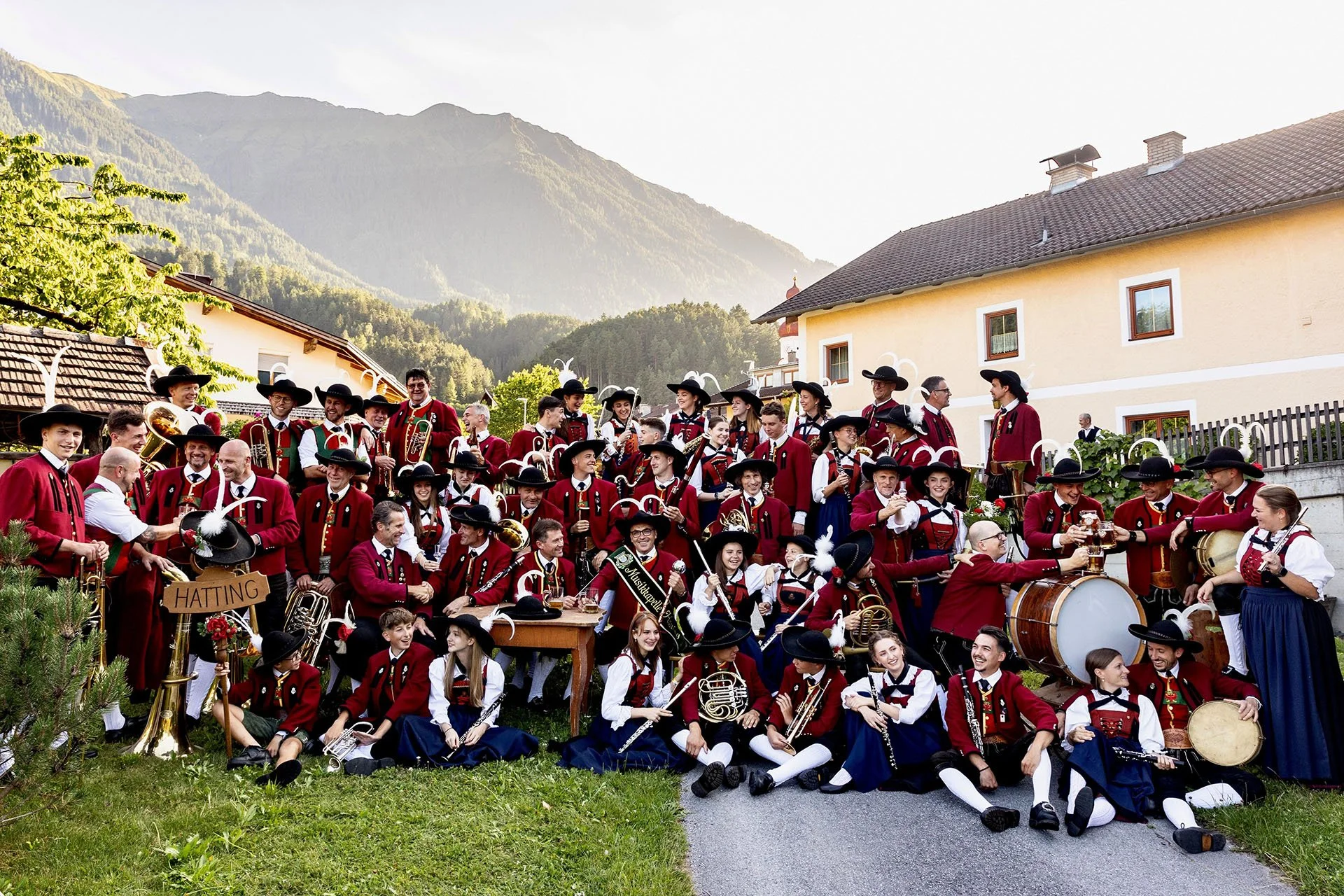 Eine große Gruppe von Menschen in traditioneller Tracht spielt Blasmusik im Freien vor Häusern und Bergen im Hintergrund.