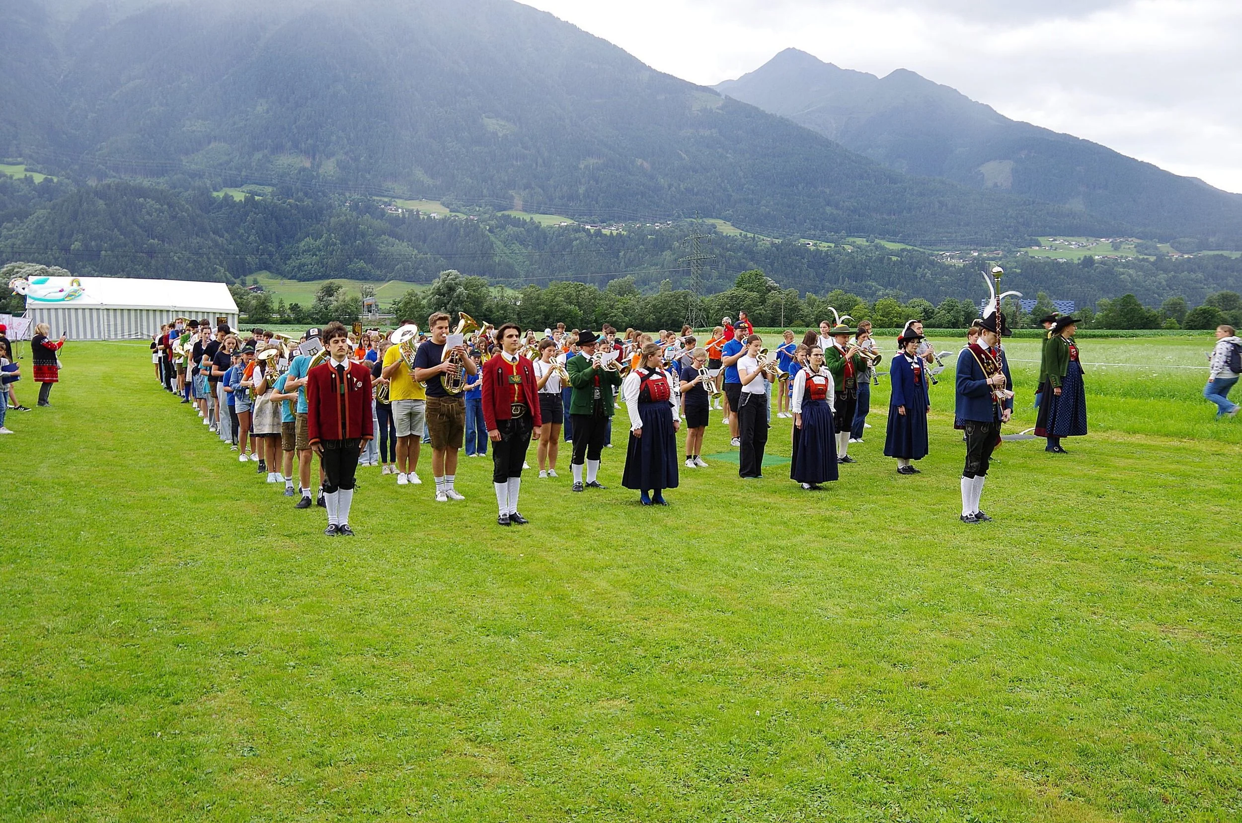 Eine Gruppe von Menschen in traditionellen Trachten, die eine Blasmusikproben im Freien auf einer grünen Wiese vor beeindruckender Bergkulisse machen.