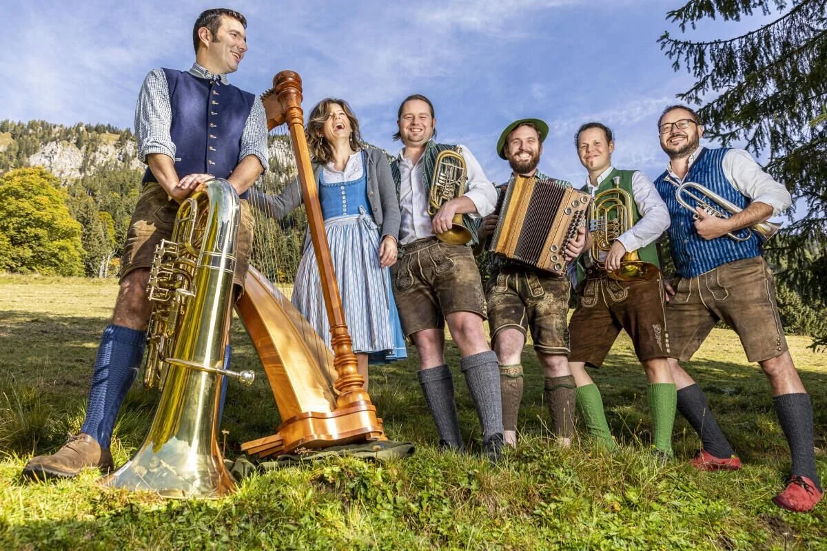 Gruppe von sieben Menschen in traditionellen bayerischen Trachten, die auf einer Wiese mit Berglandschaft im Hintergrund stehen, einige halten Musikinstrumente wie Tuba, Harfe, Akkordeon und Trompete, beim gemeinsamen Musizieren an sonnigem Tag.