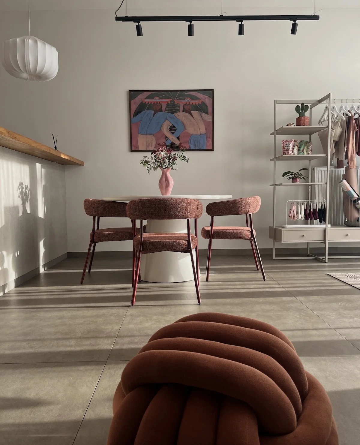Modern dining room with a round table and three pink upholstered chairs, pink vase with flowers on the table, abstract wall art above, white shelving unit with plants and clothing, sunlight casting shadows.