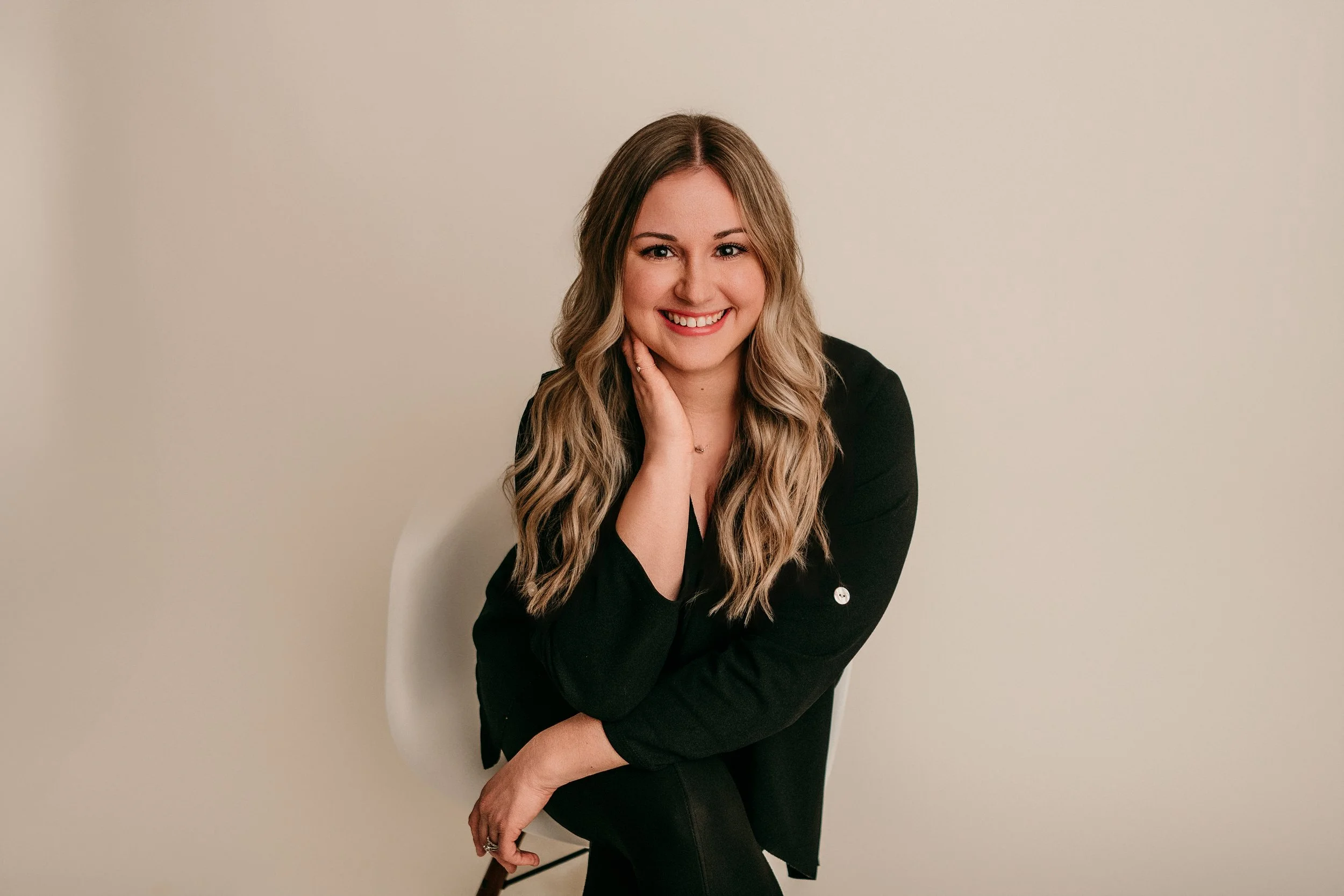 A woman with long blonde hair, wearing a black blazer, smiling and sitting on a chair against a plain beige wall.