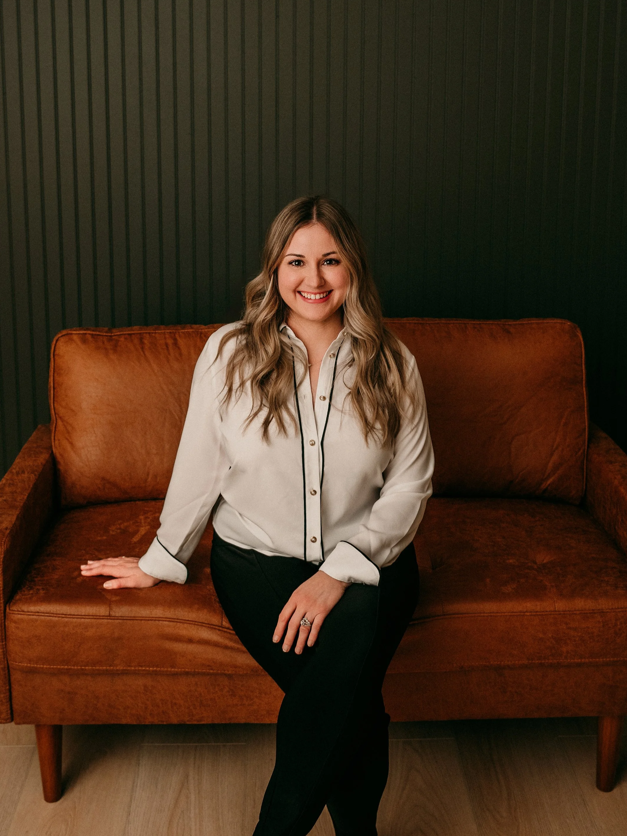A woman with long wavy blonde hair sitting on a brown leather sofa against a dark panel wall, smiling at the camera.