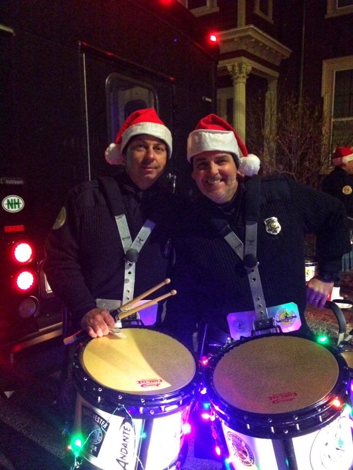 Two men wearing Santa hats and police uniforms standing next to a drum set with Christmas lights, smiling at the camera.