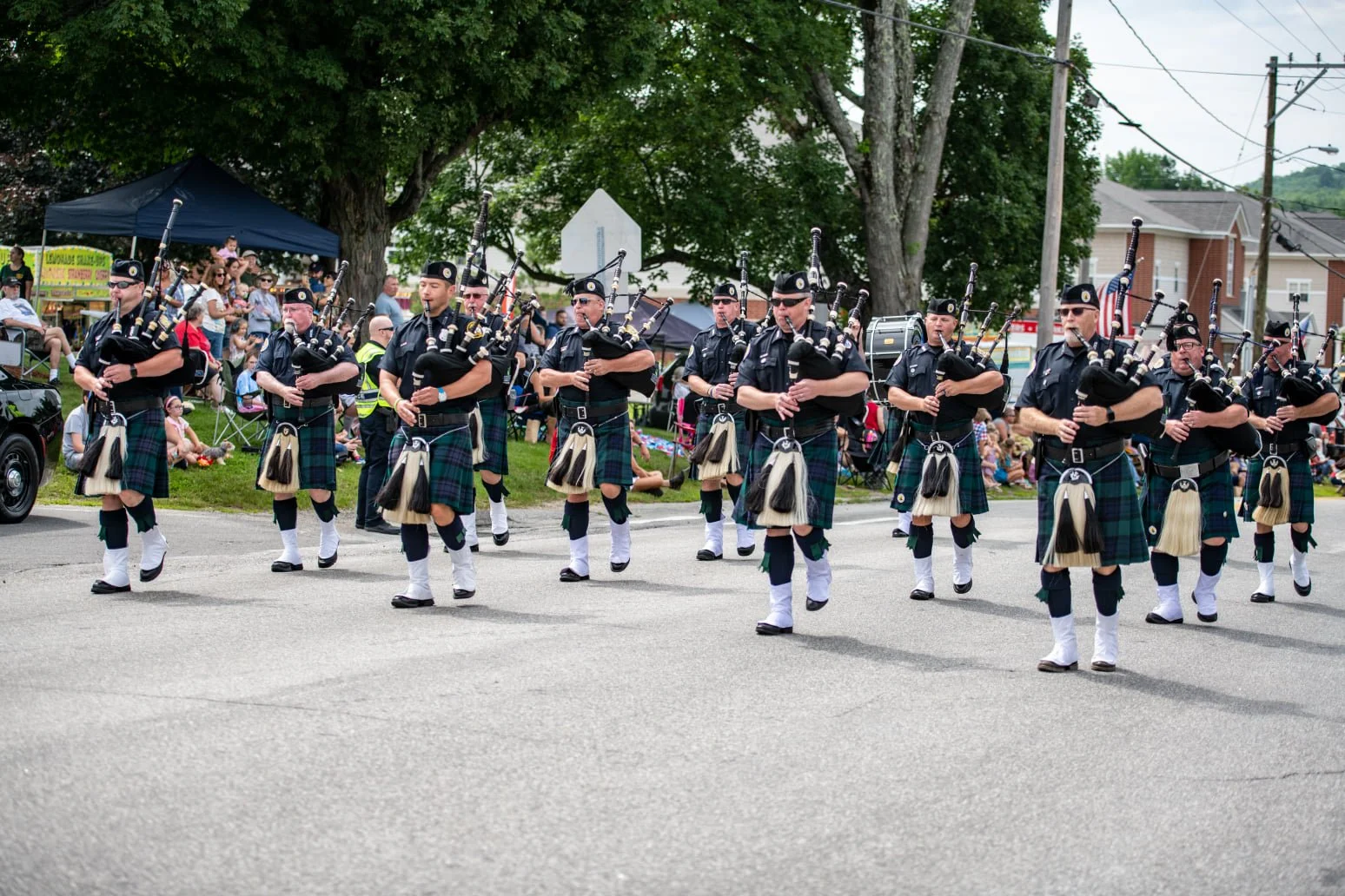 A parade of bagpipers dressed in traditional Scottish attire marching on a street during a festive event with spectators watching from the sidelines.