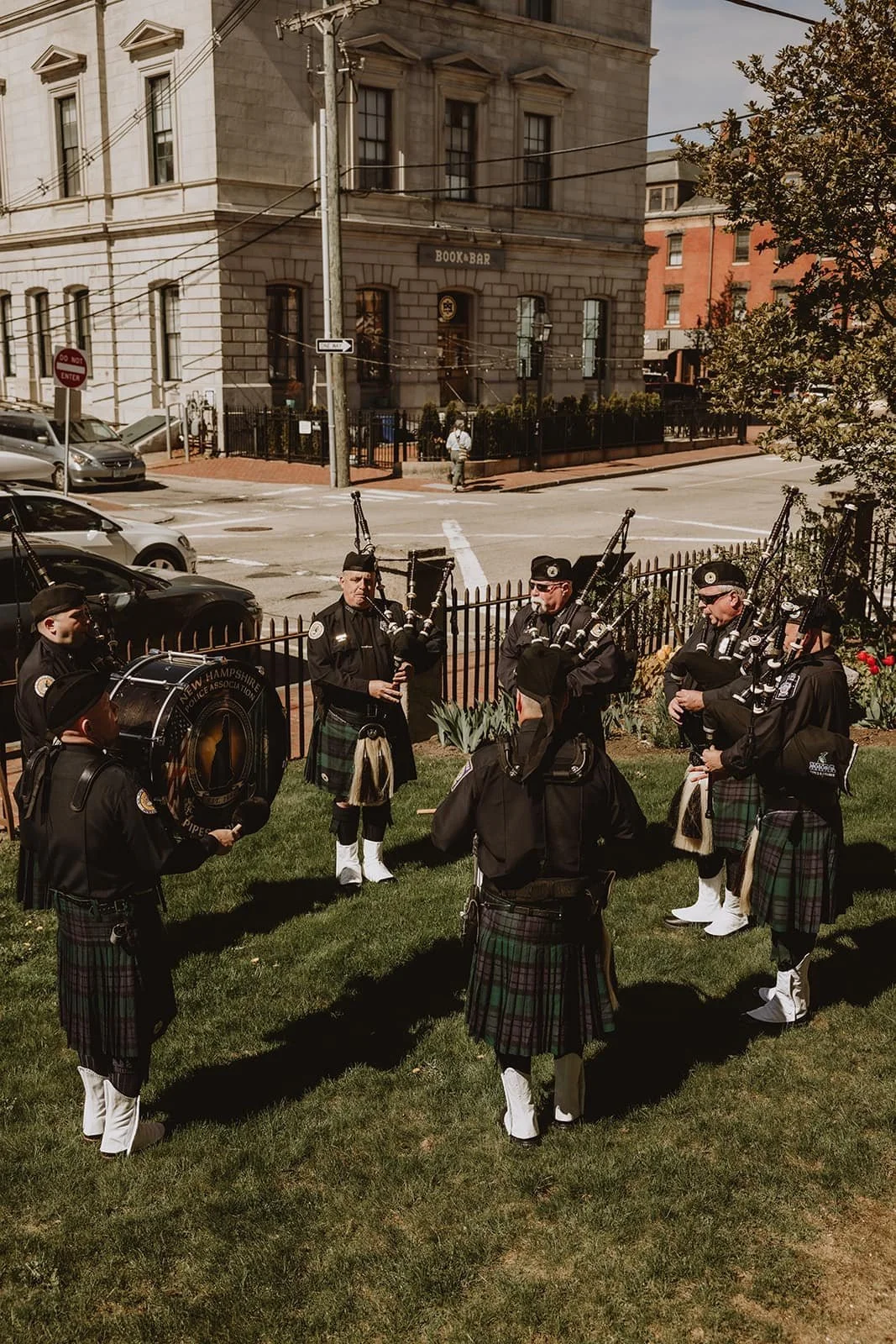 A group of seven police officers in traditional Scottish kilts playing bagpipes and a drum on a small patch of grass near a fence on a city street.